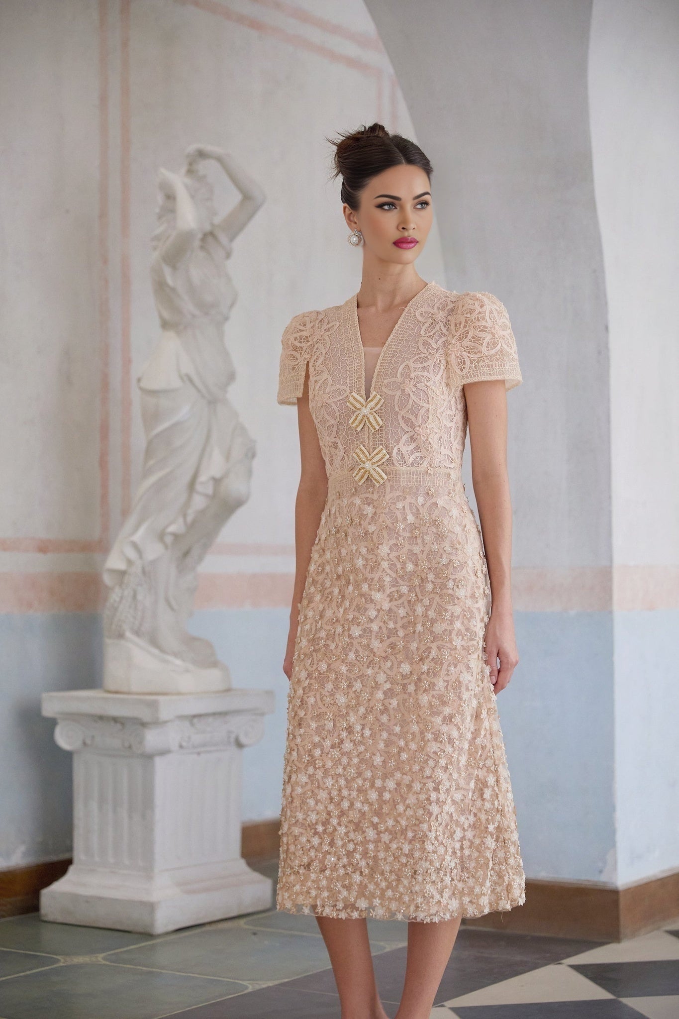 A beige midi dress with a crochet pattern and faux flower details worn by a model standing in a neutral setting