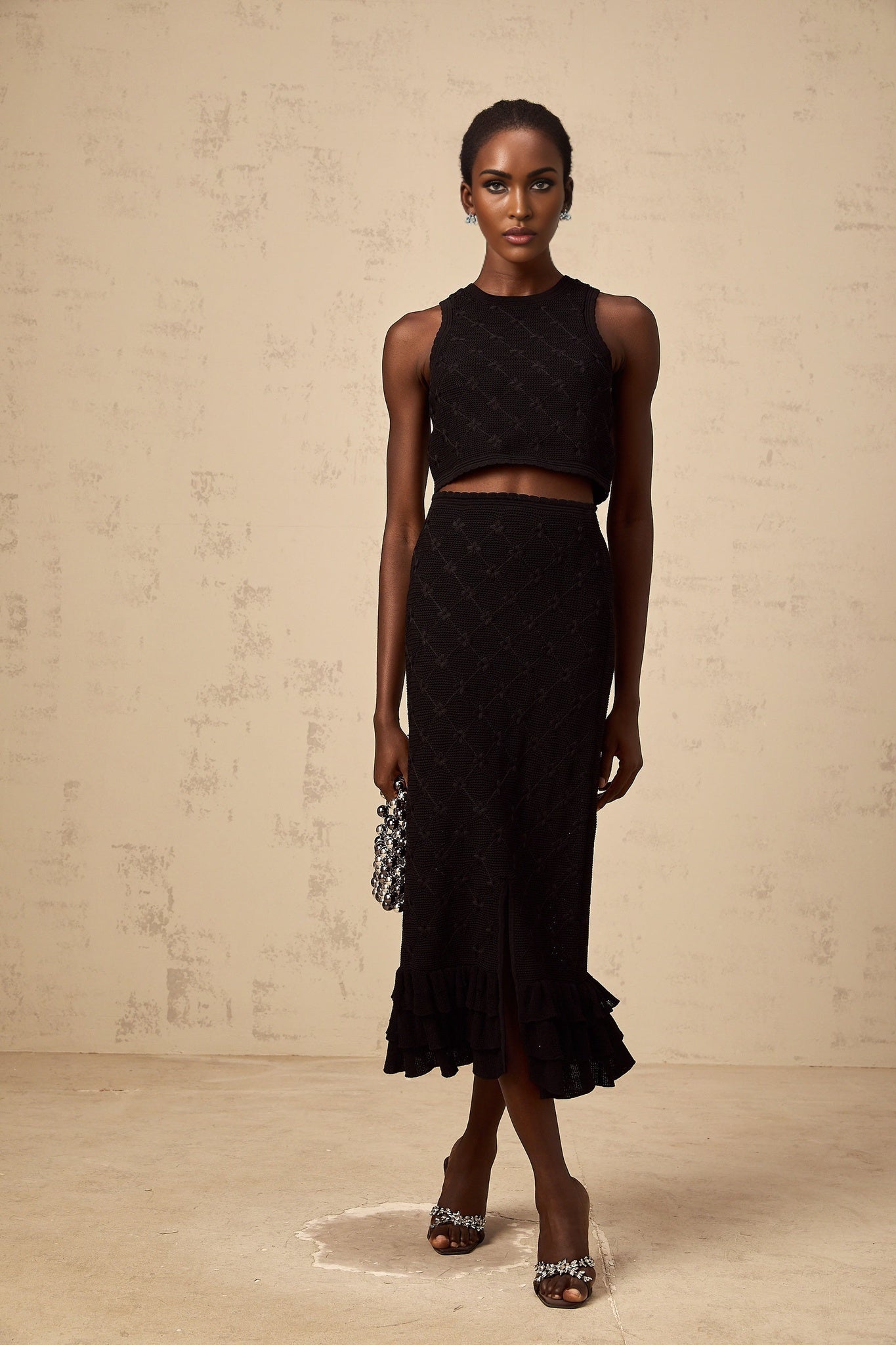 A black crochet knit matching set consisting of a top and skirt worn by a model against a plain background