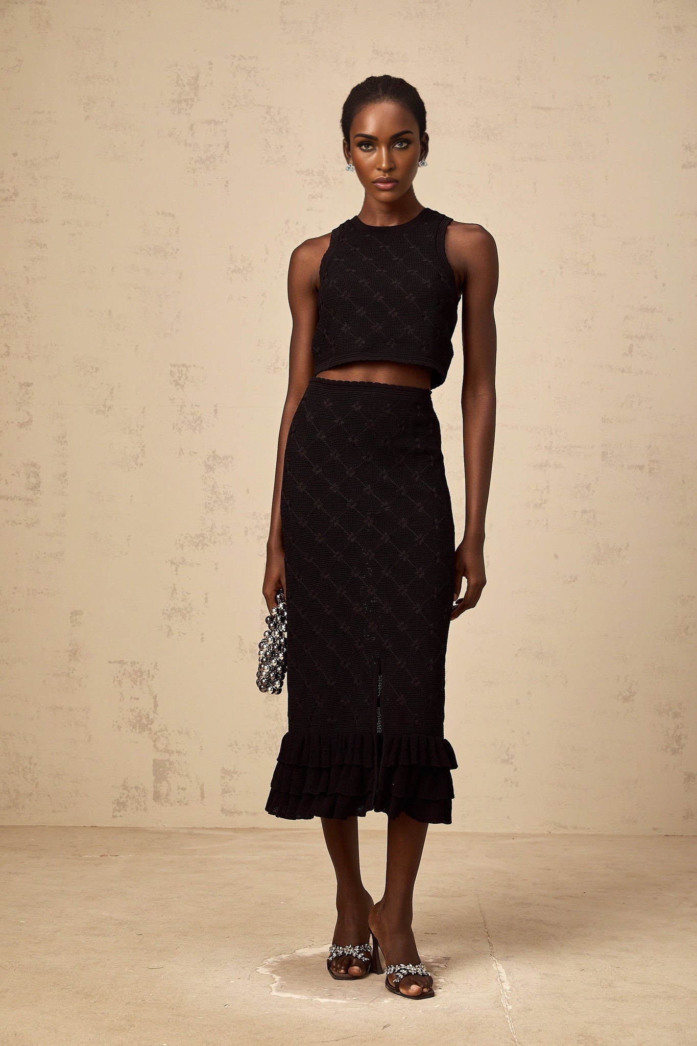 A black crochet knit matching set consisting of a top and skirt worn by a model against a plain background