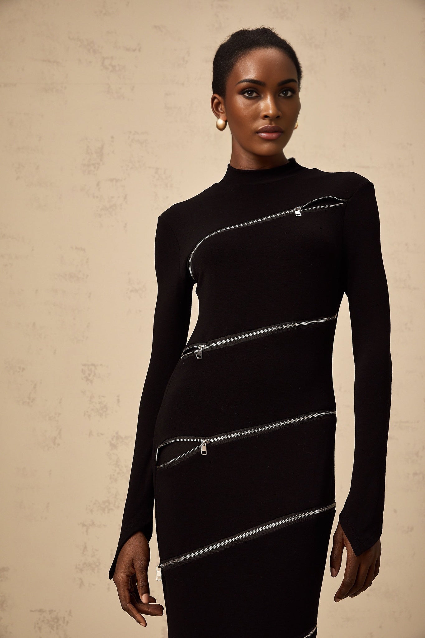 A black fitted maxi dress with zipper details worn by a model standing against a plain background