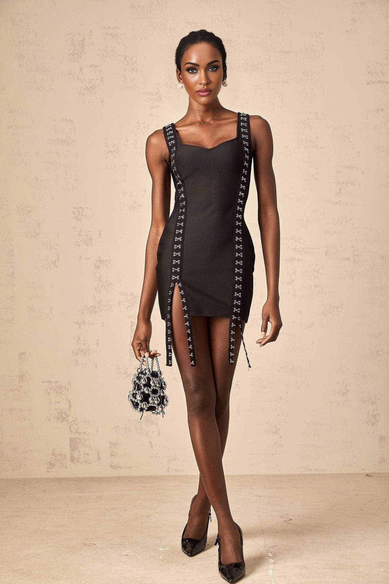 A black formfitting mini dress with hookandeye closures worn by a model standing against a plain background