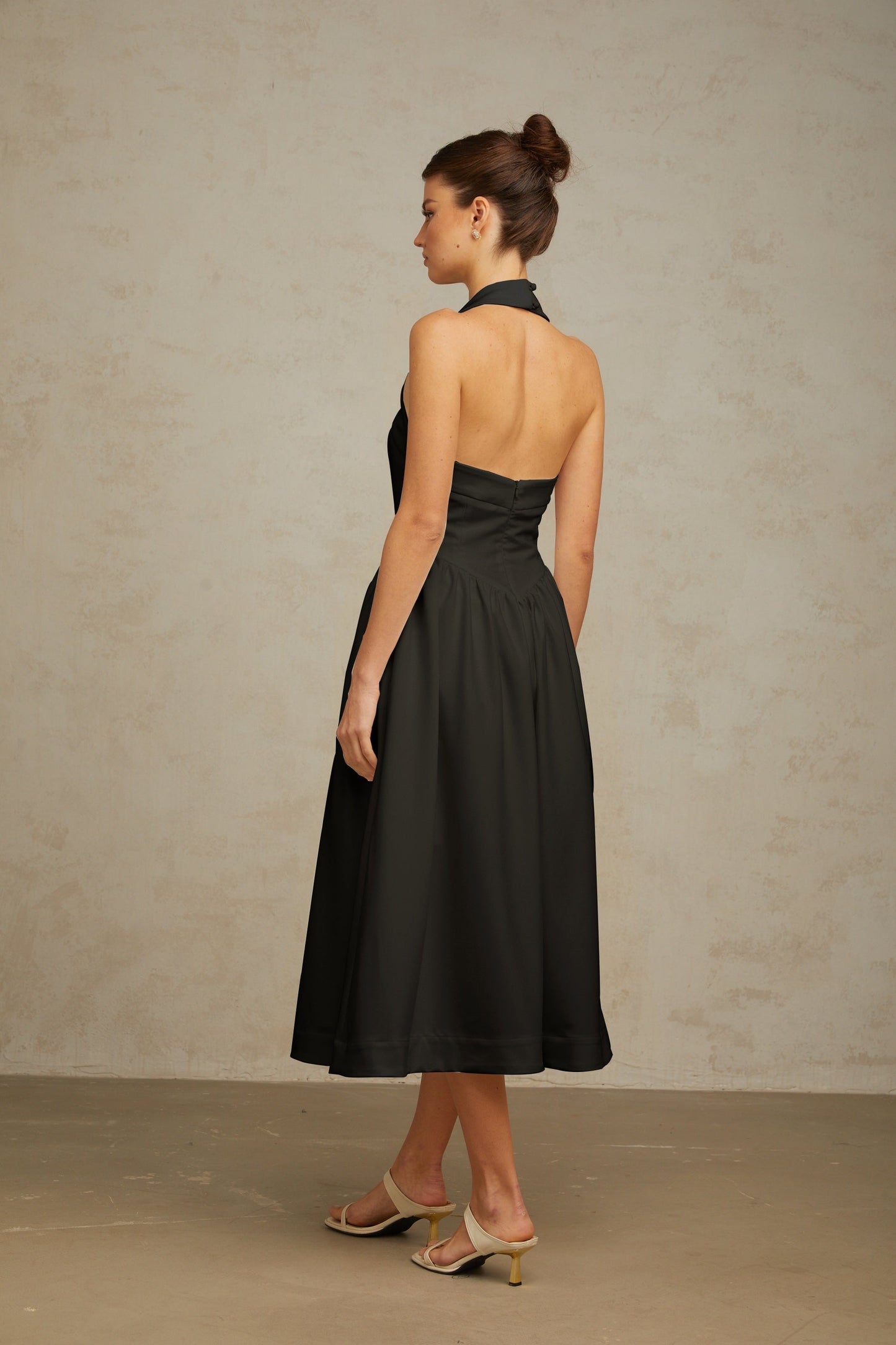 A black halterneck midi dress with pleats and a flowy silhouette worn by a model standing against a plain background