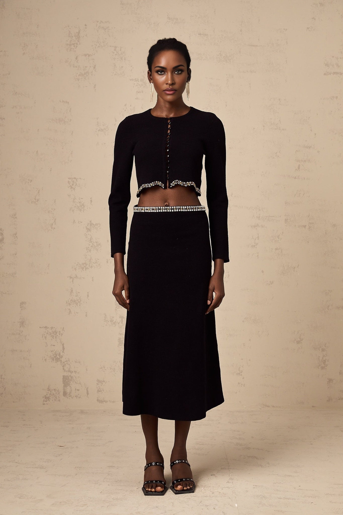 A black knitted cardigan and skirt set embellished with decorative details worn by a model showing the matching outfit