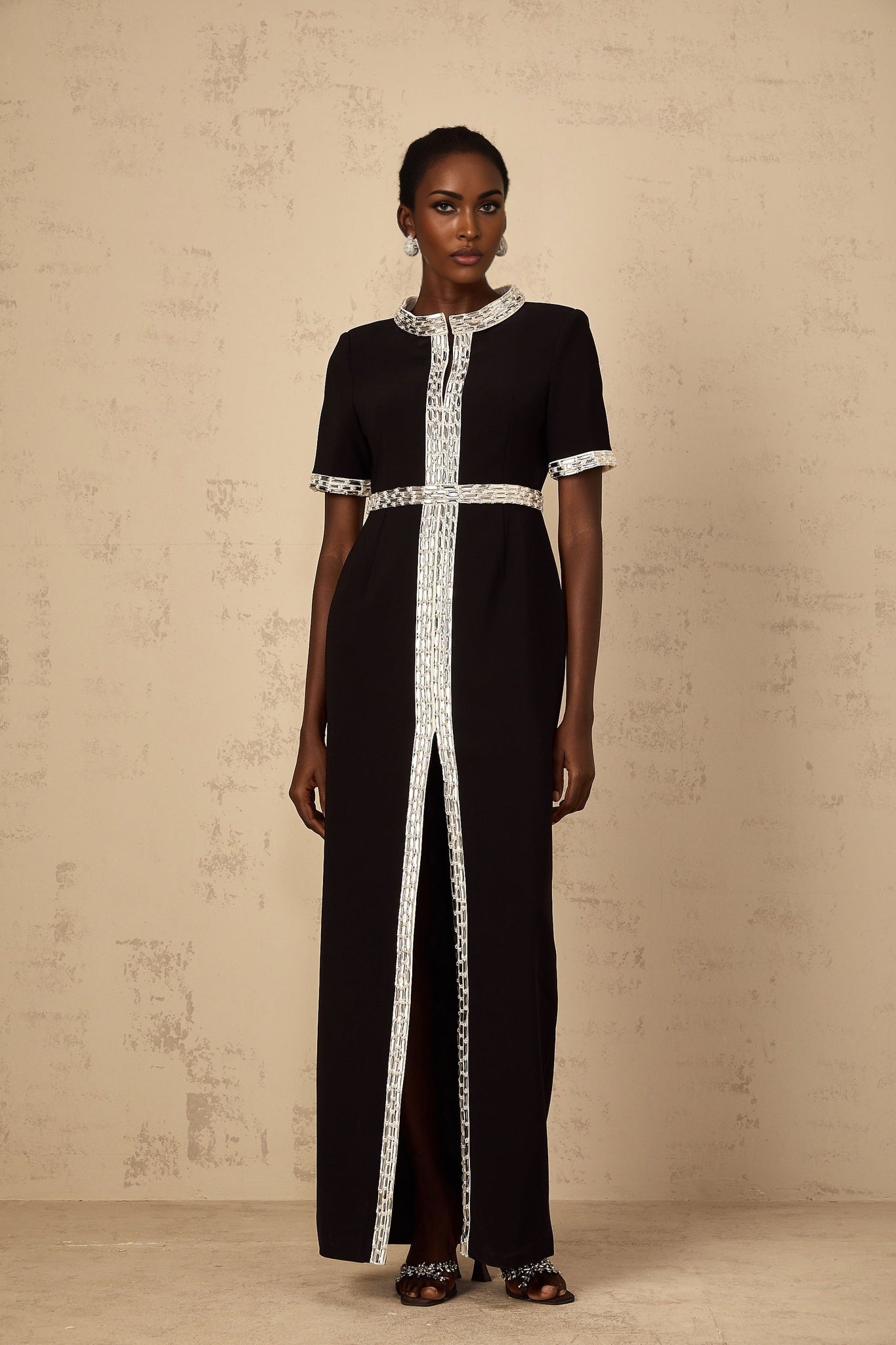 A black maxi dress with a front slit and crystal embellishments worn by a model standing against a plain background
