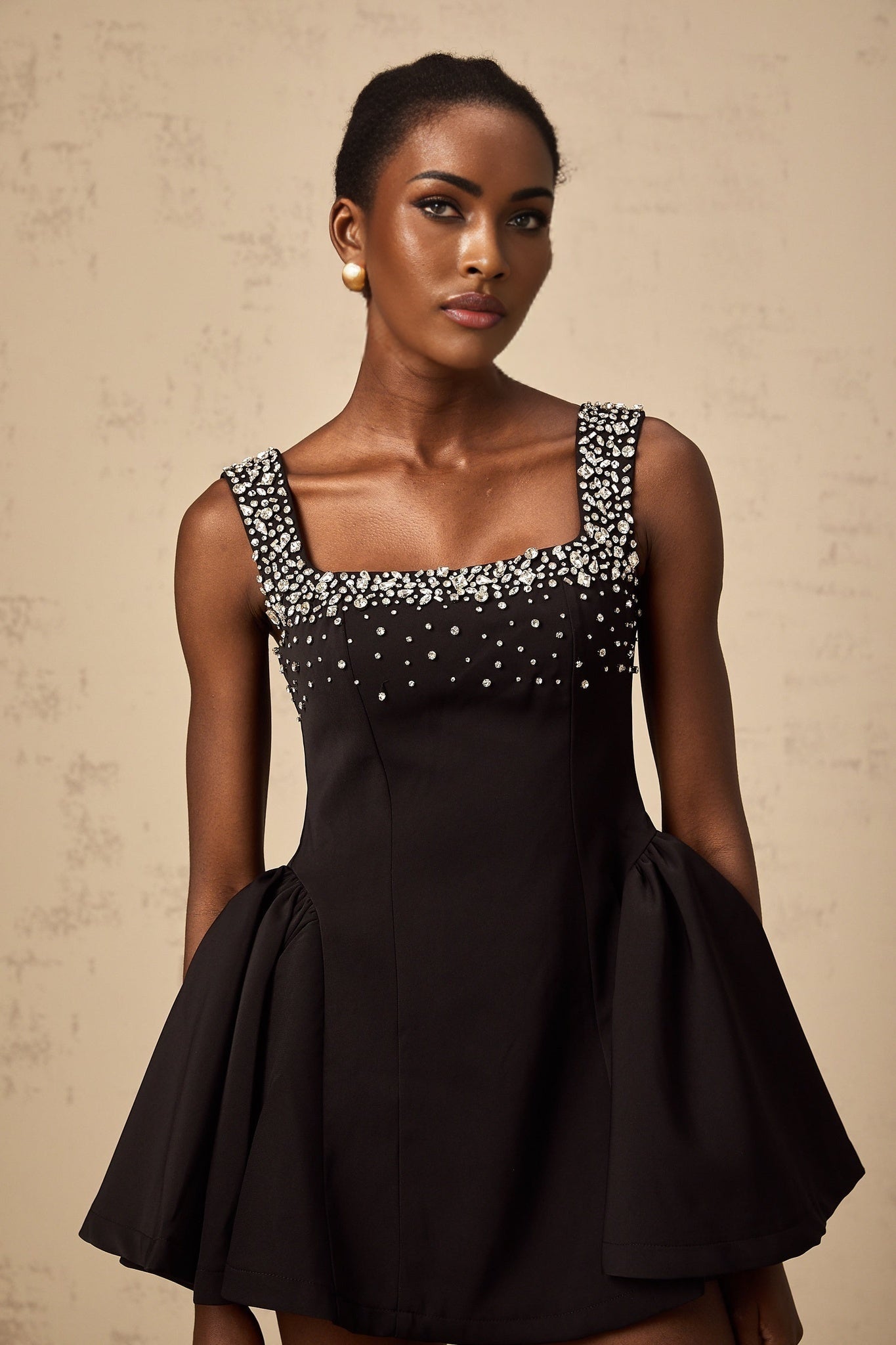 A black mini dress with a flared skirt adorned with crystal embellishments worn by a model standing against a plain background