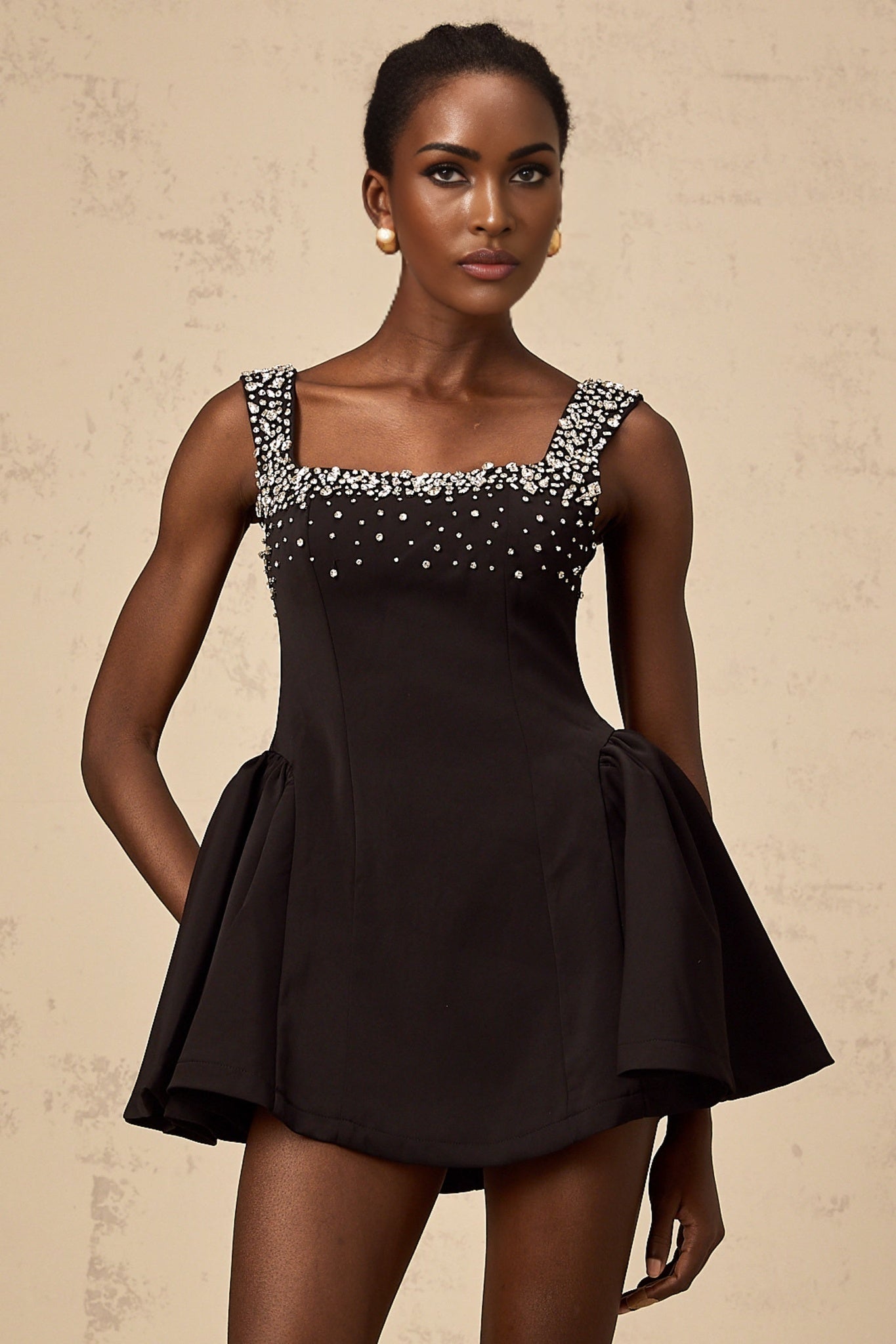 A black mini dress with a flared skirt featuring crystal embellishments worn by a model standing against a plain background