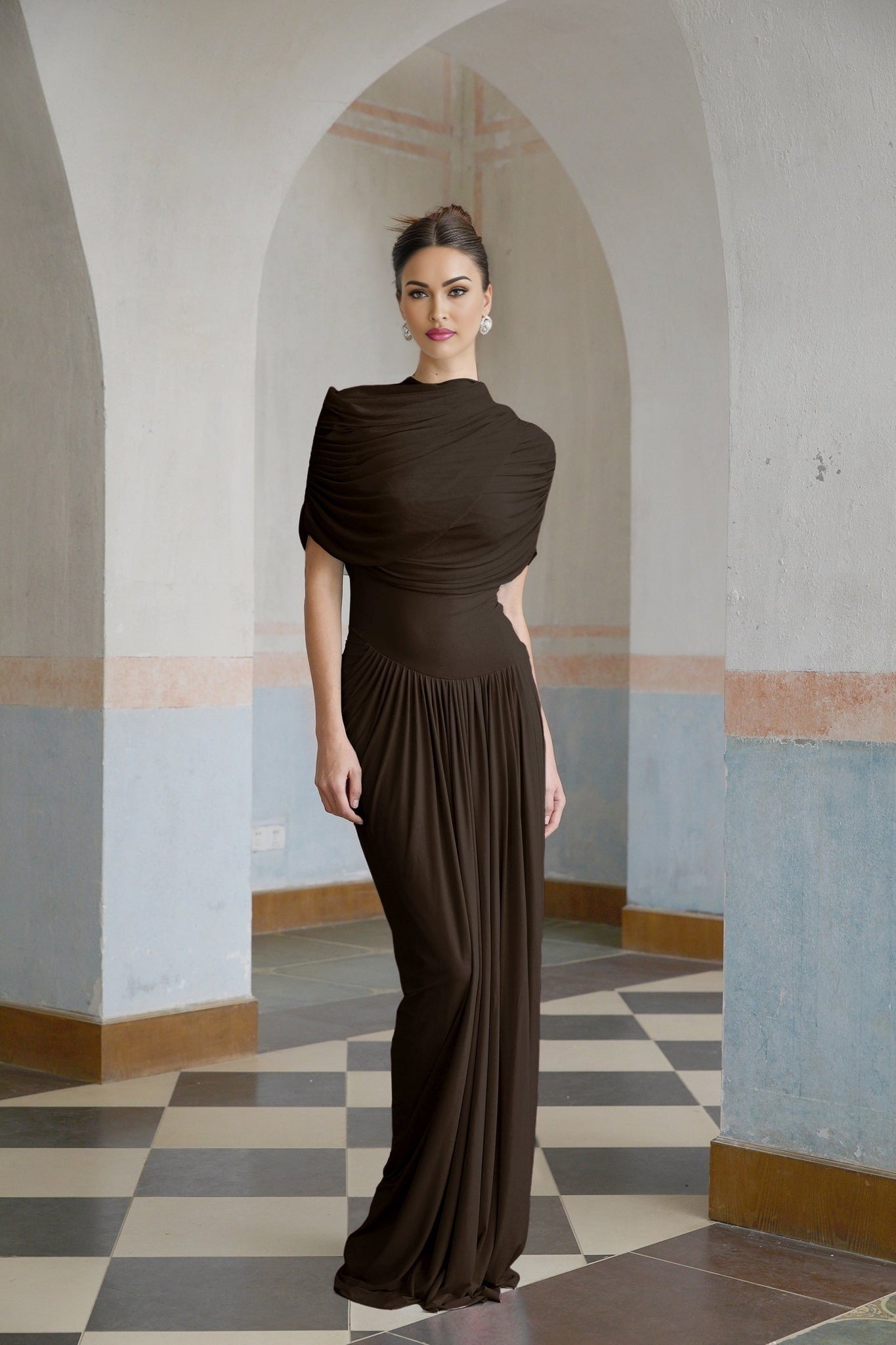 A brown asymmetric maxi dress with pleated design and flowing skirt worn by a female model standing against a plain background