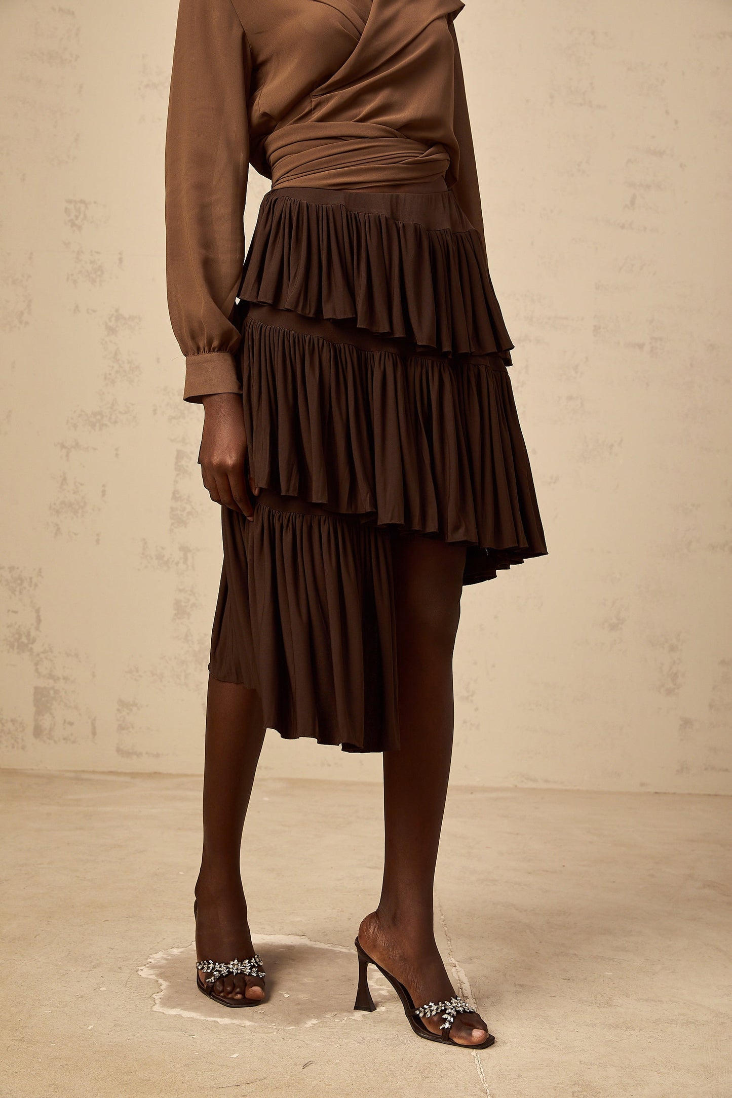 A brown asymmetrical mini skirt with pleated tiers and a unique layered design worn by a model against a plain background