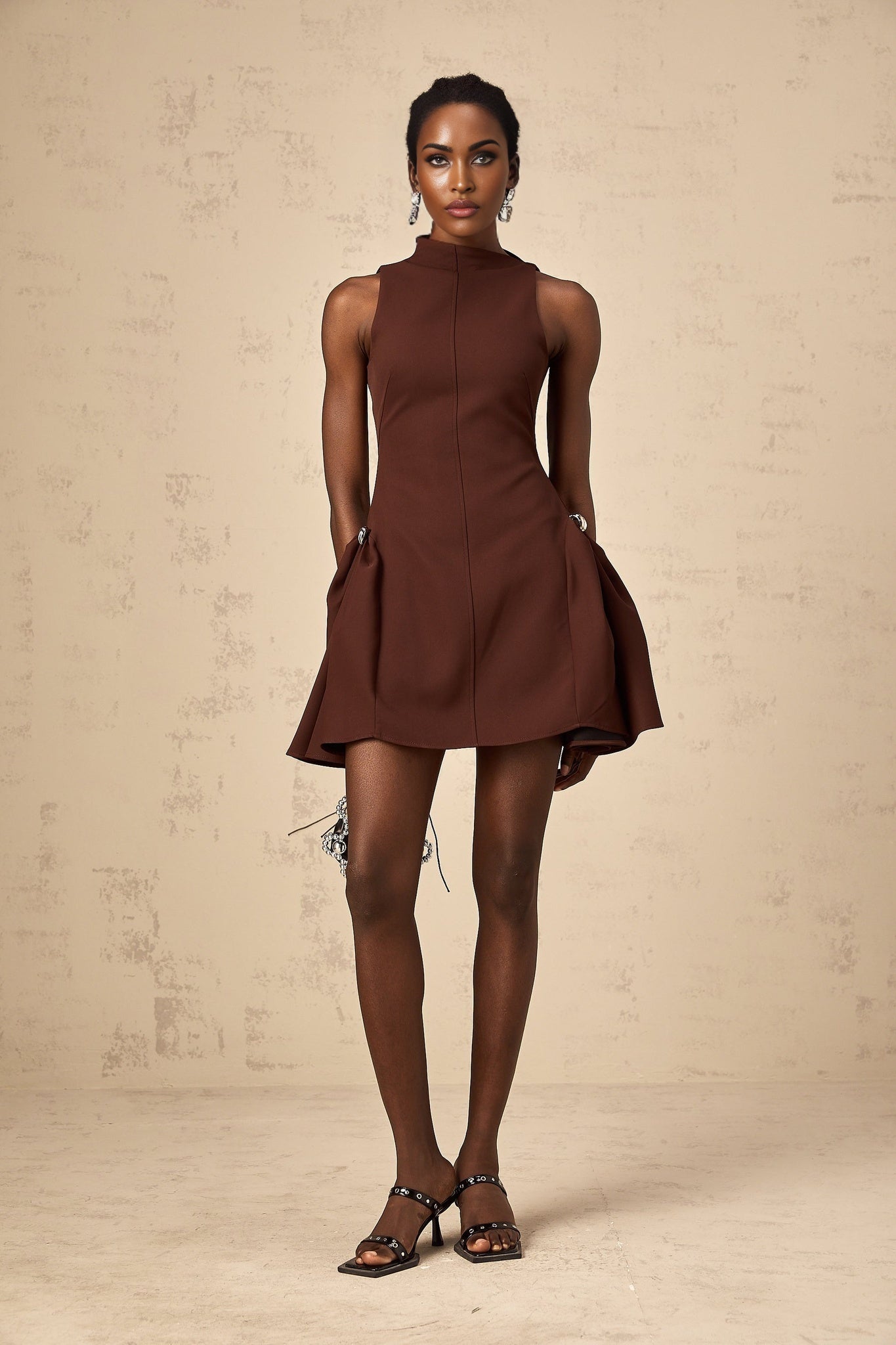 A brown mini dress with an asymmetric neckline and puff sleeves worn by a model named Dora standing against a plain background