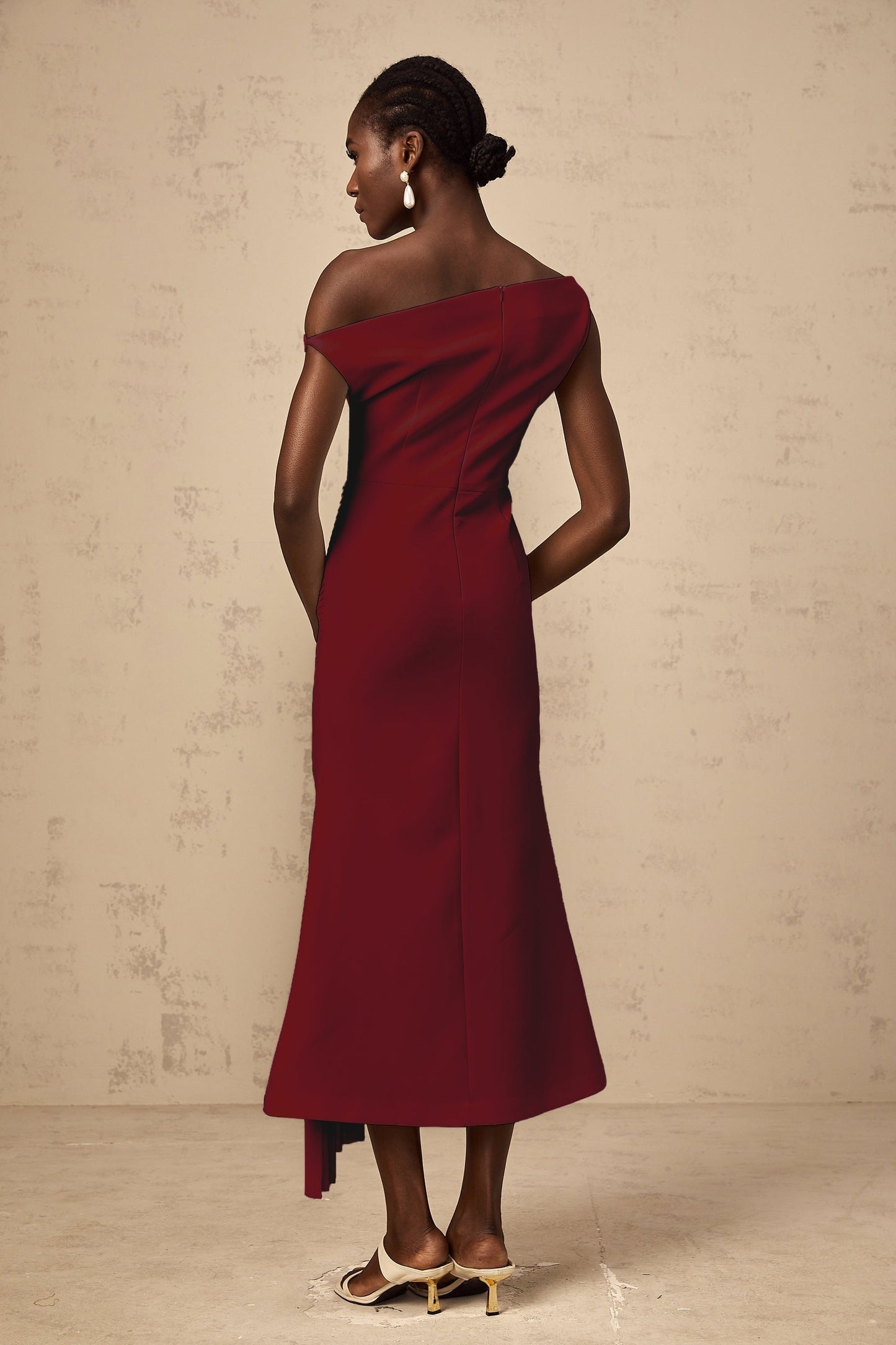 A burgundy colored asymmetrical midi dress with draped details worn by a model standing against a plain background