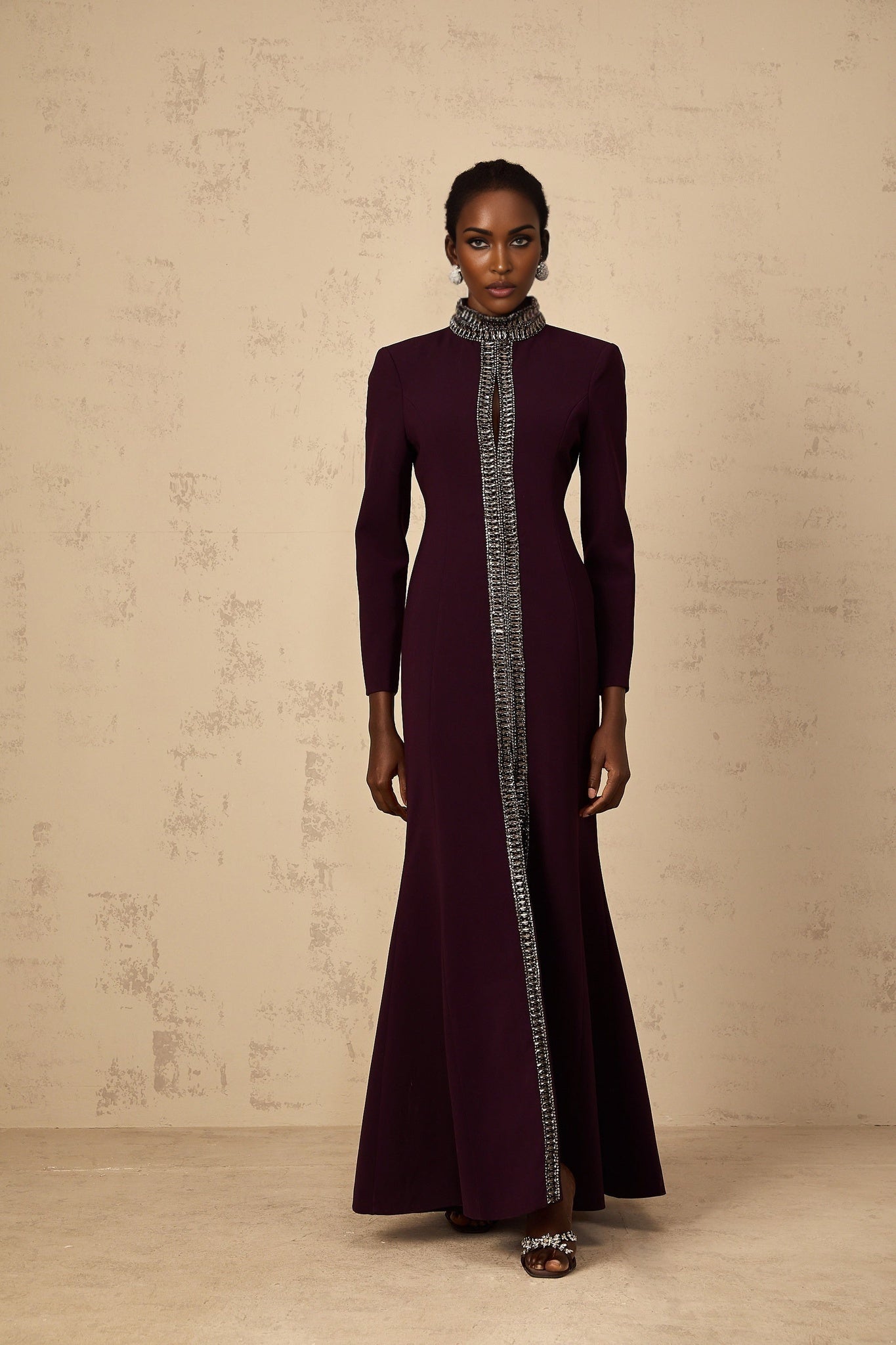 A burgundy maxi dress with a flared skirt and embellished details worn by a model standing against a plain background