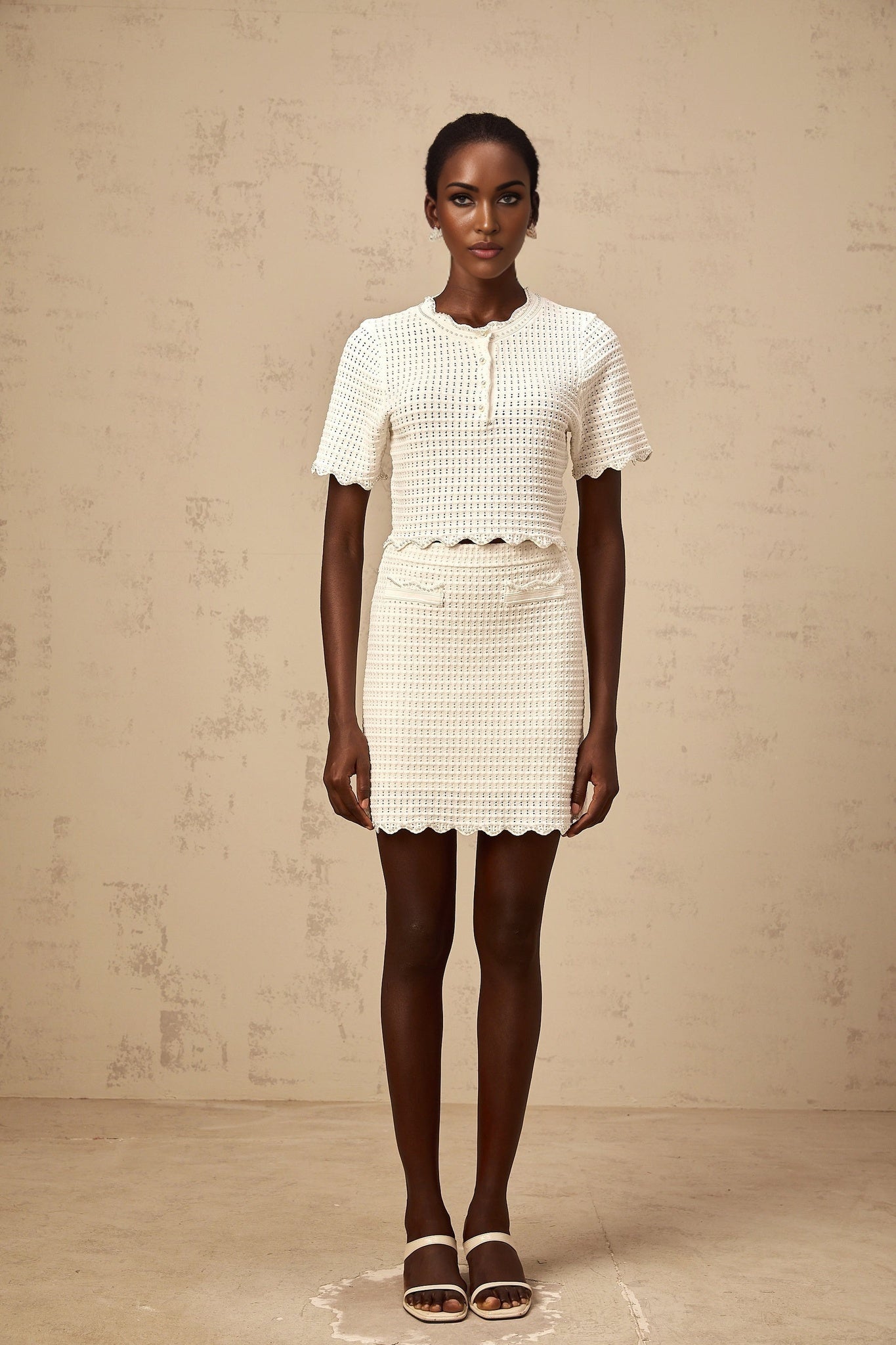 A creamwhite matching set featuring a ruffled top and skirt made of pointelle knit fabric The top has a delicate ruffled design while the skirt complements it with a similar texture Both pieces are lightweight and have an intricate knit pattern