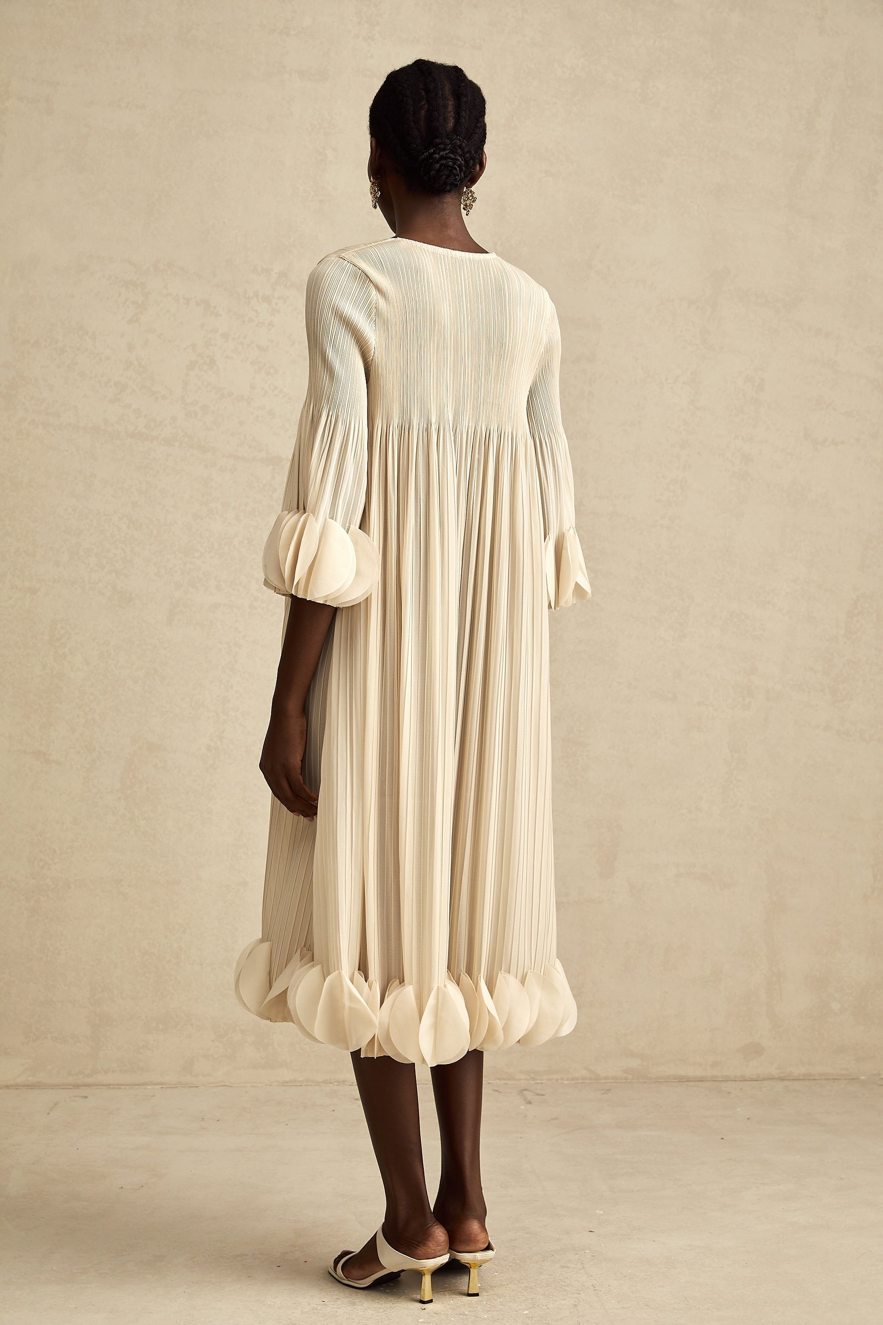 A female model wearing a midi length dress with three dimensional petal pleated details throughout the garment in a soft flowing fabric