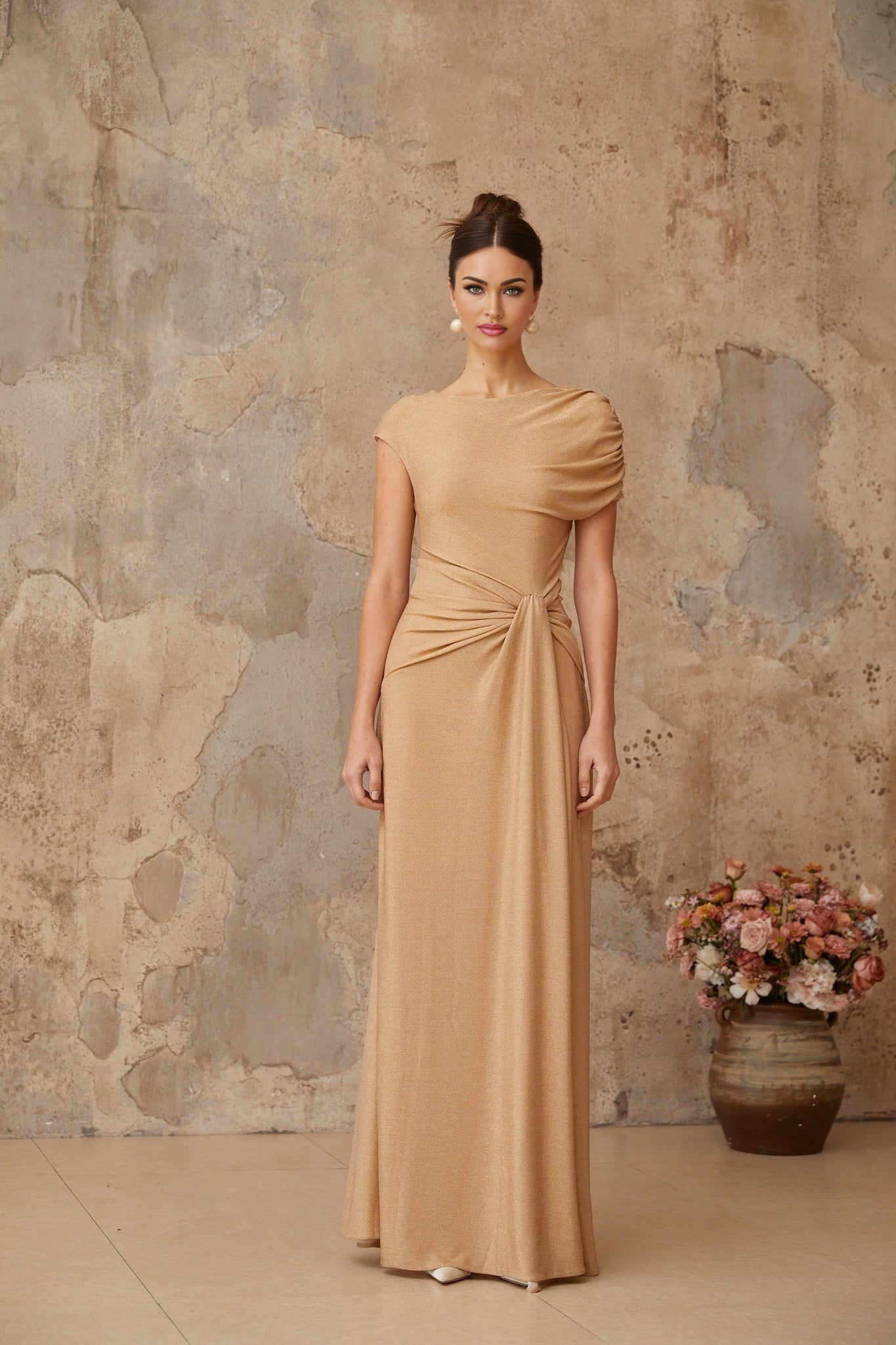 A gold maxi dress with an asymmetrical design featuring pleated fabric and a long flowing skirt The dress has a sleek modern look with a unique neckline and draping details