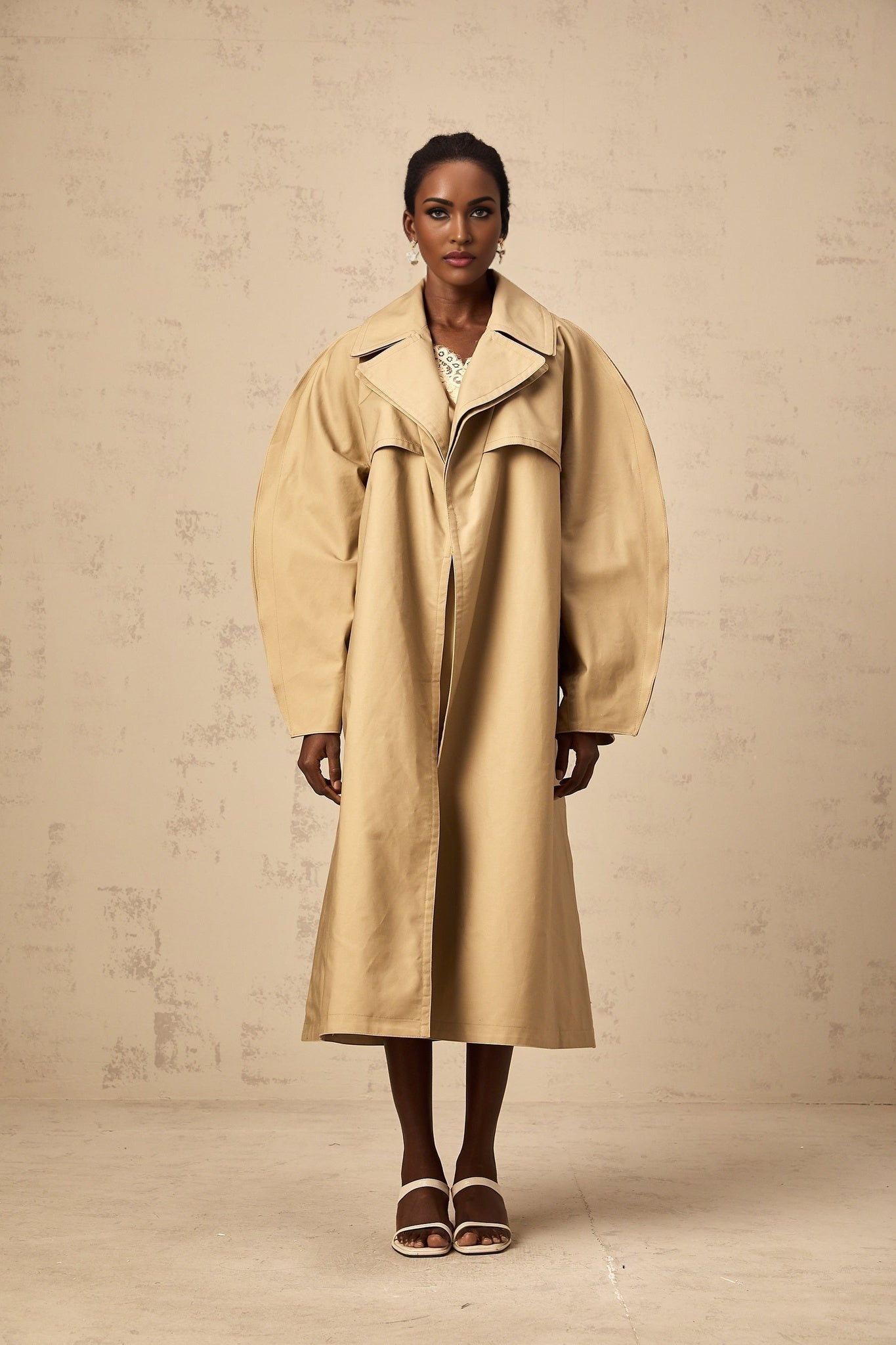 A khaki trench coat with curved sleeves worn by a model featuring a belted waist and classic lapels