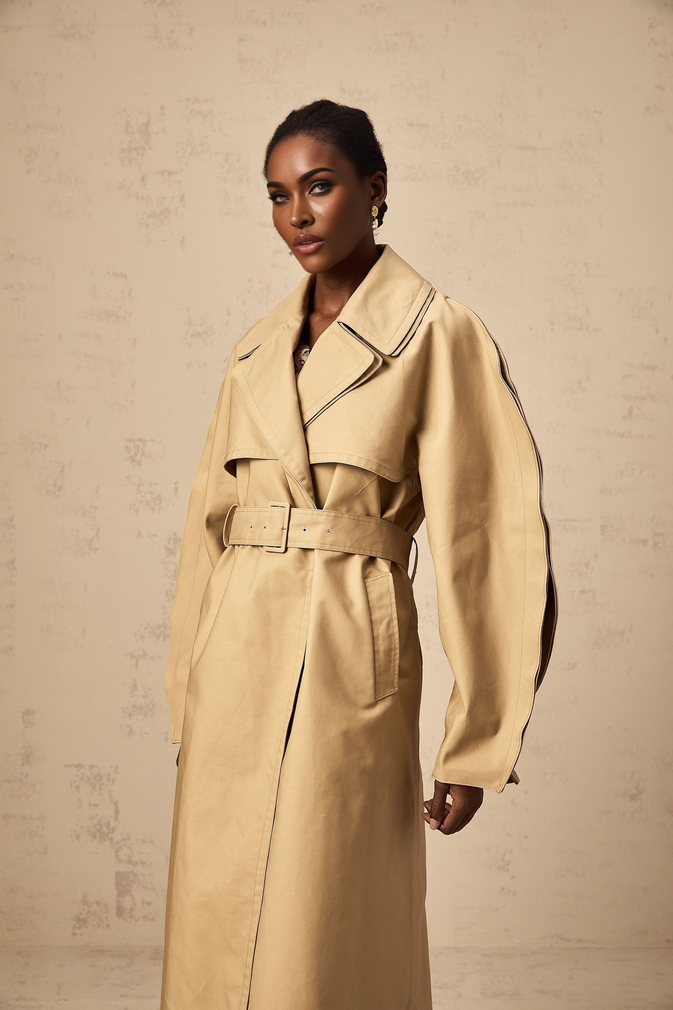 A khaki trench coat with curved sleeves worn by a model standing against a plain background The coat features a classic doublebreasted design with a belt at the waist and epaulettes on the shoulders