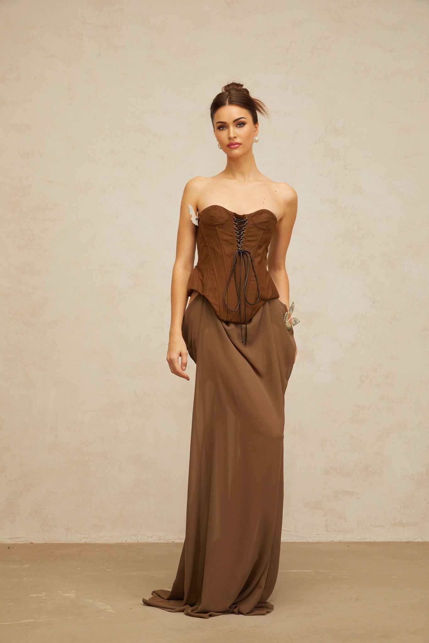 A long brown maxi dress with corsetstyle bodice featuring butterflyshaped pin details and a flowing skirt