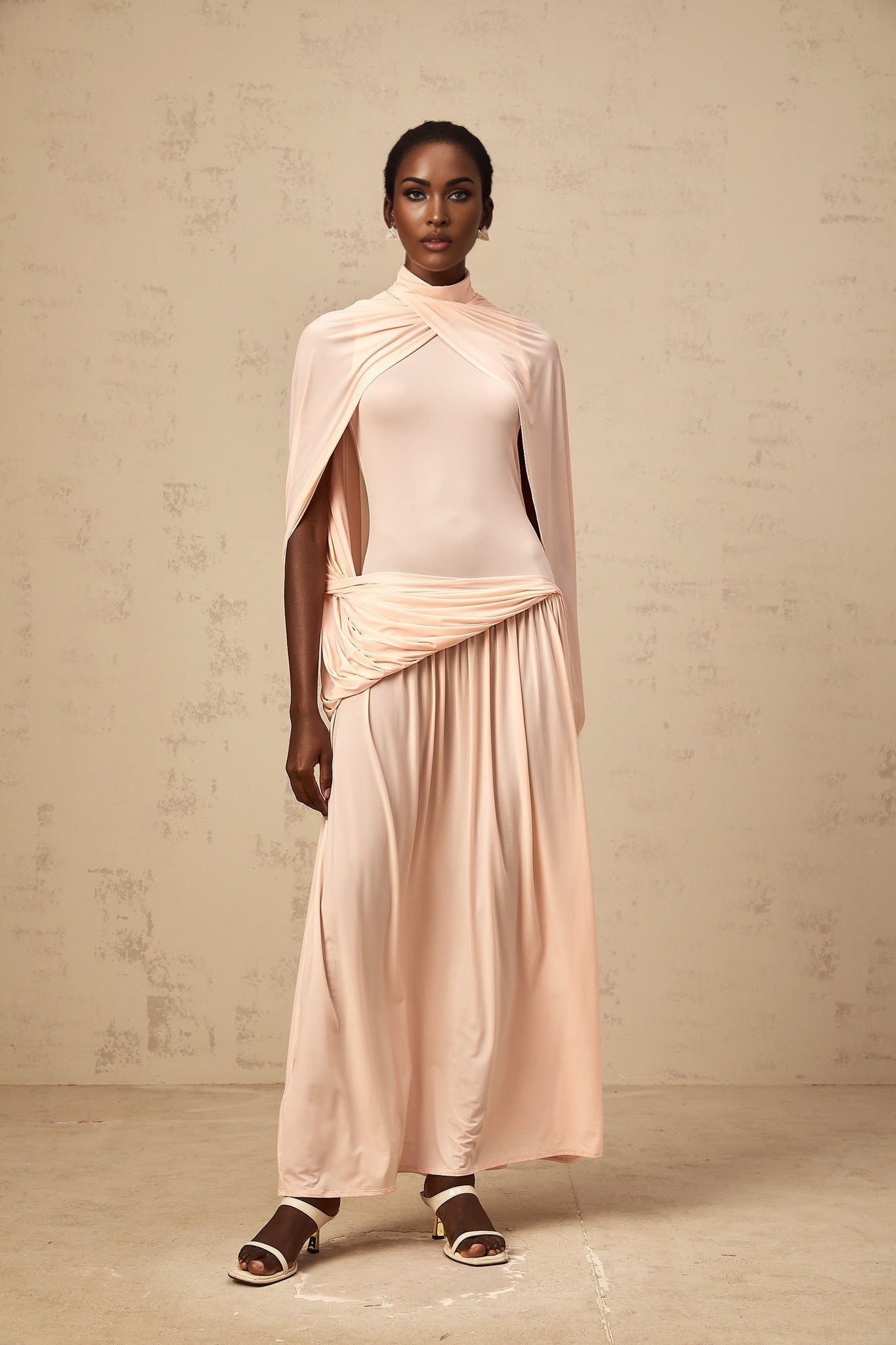 A long flowing pink maxi dress with an asymmetrical hemline and a capelike design over the shoulders and upper body