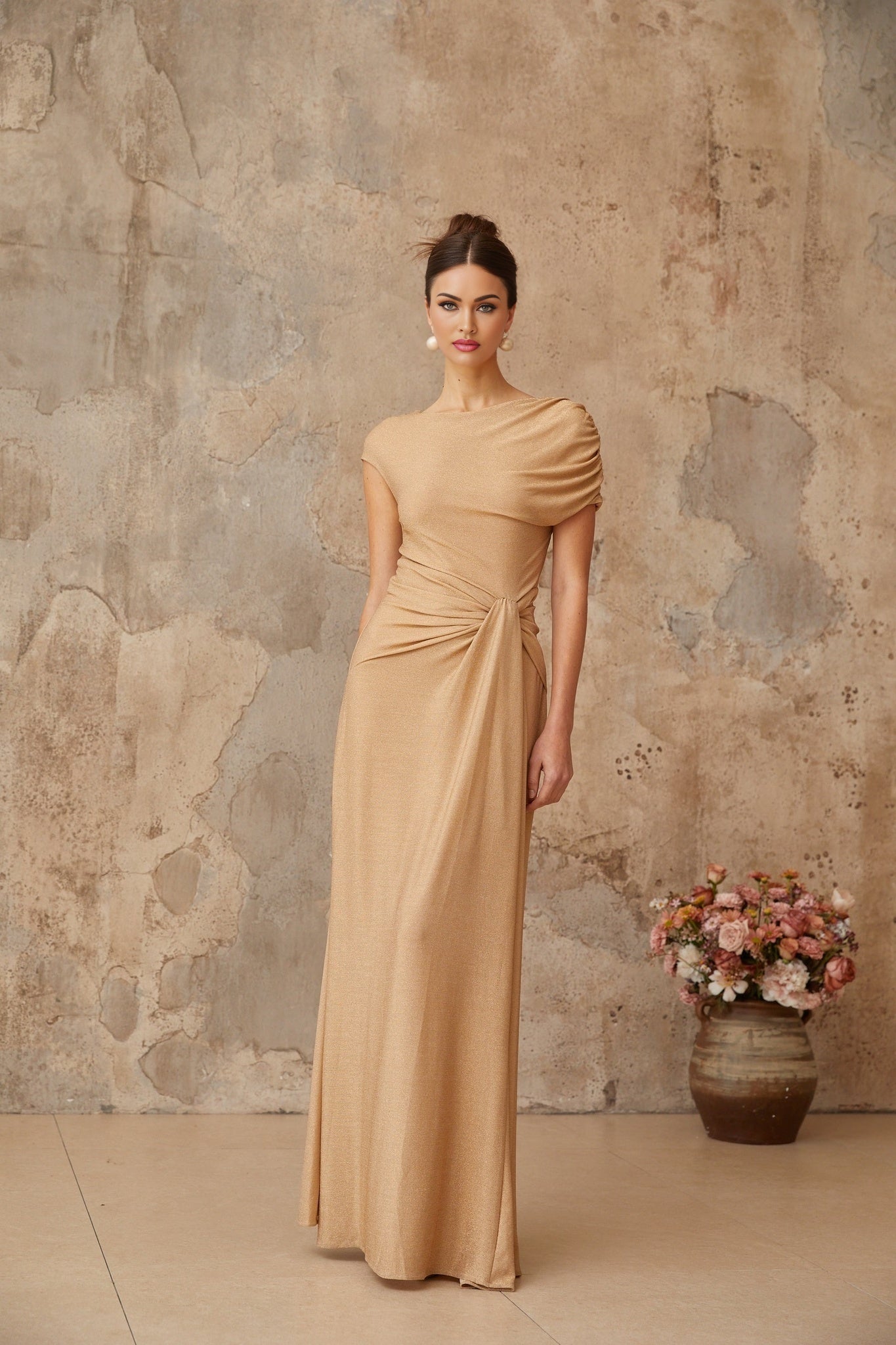 A long gold maxi dress with an asymmetrical hem and pleated design featuring a flowy silhouette and shimmering fabric