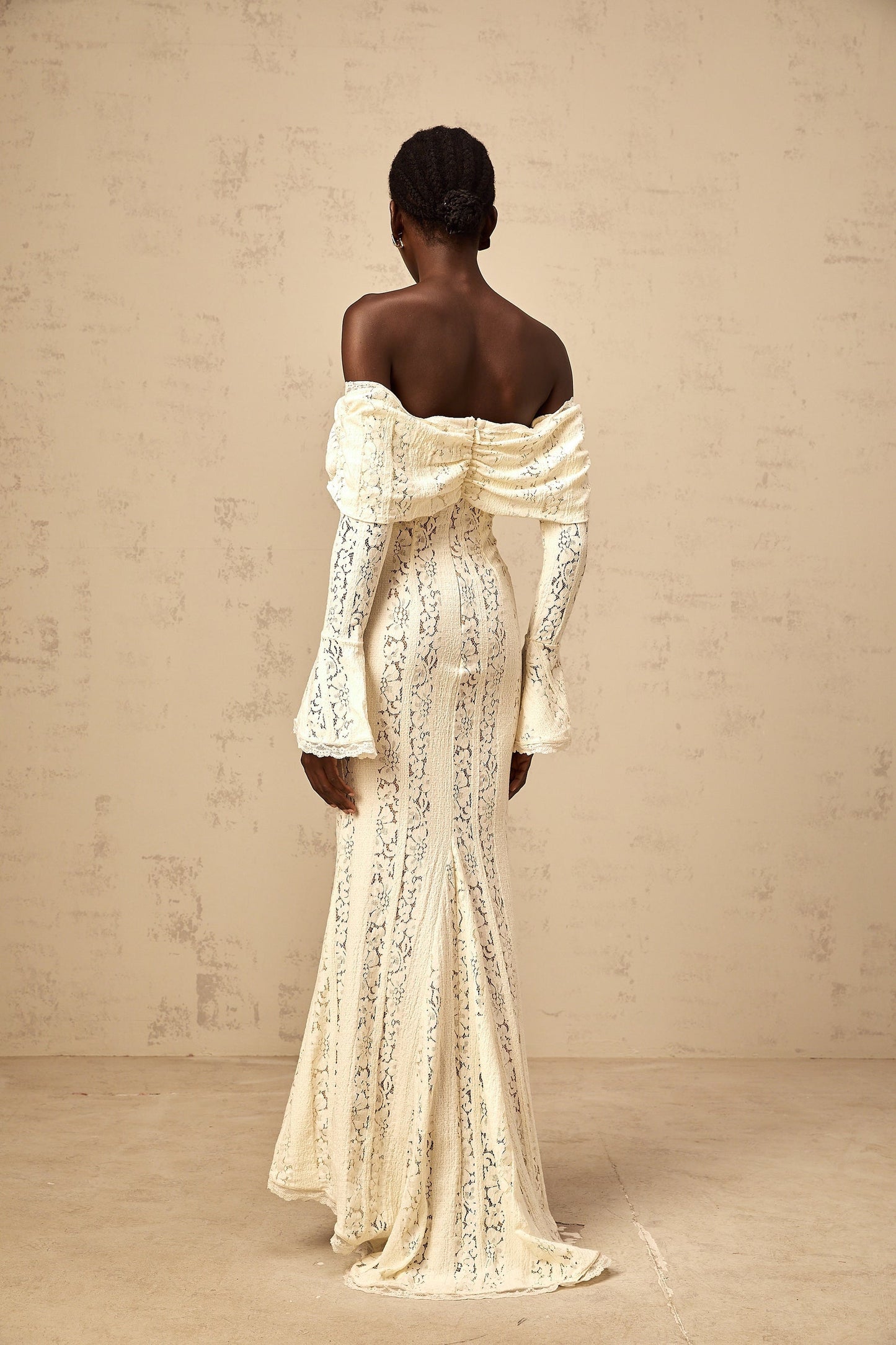 A long neutral colored maxi dress with floral lace detailing and a train design worn by a model standing in a studio setting