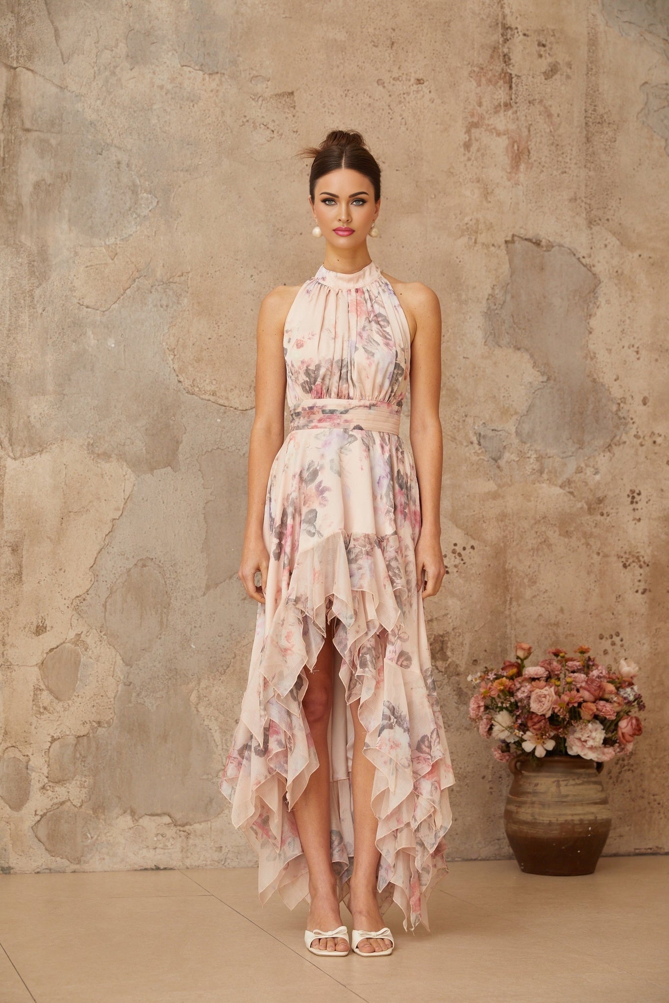 A long pink maxi dress with an asymmetrical hemline and allover floral print pattern worn by a model standing against a plain background