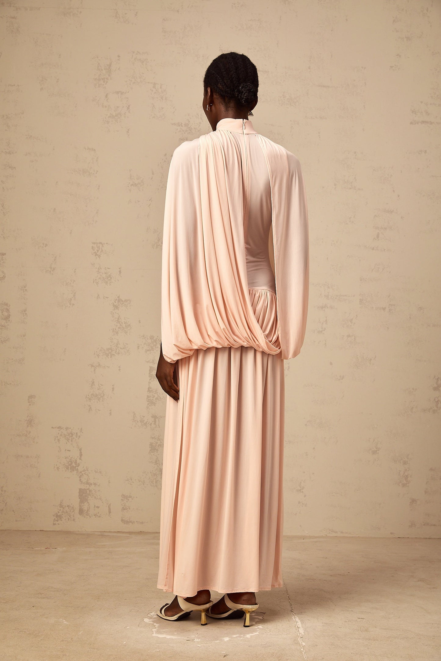 A long pink maxi dress with an asymmetrical hemline featuring a flowing cape design draped over one shoulder