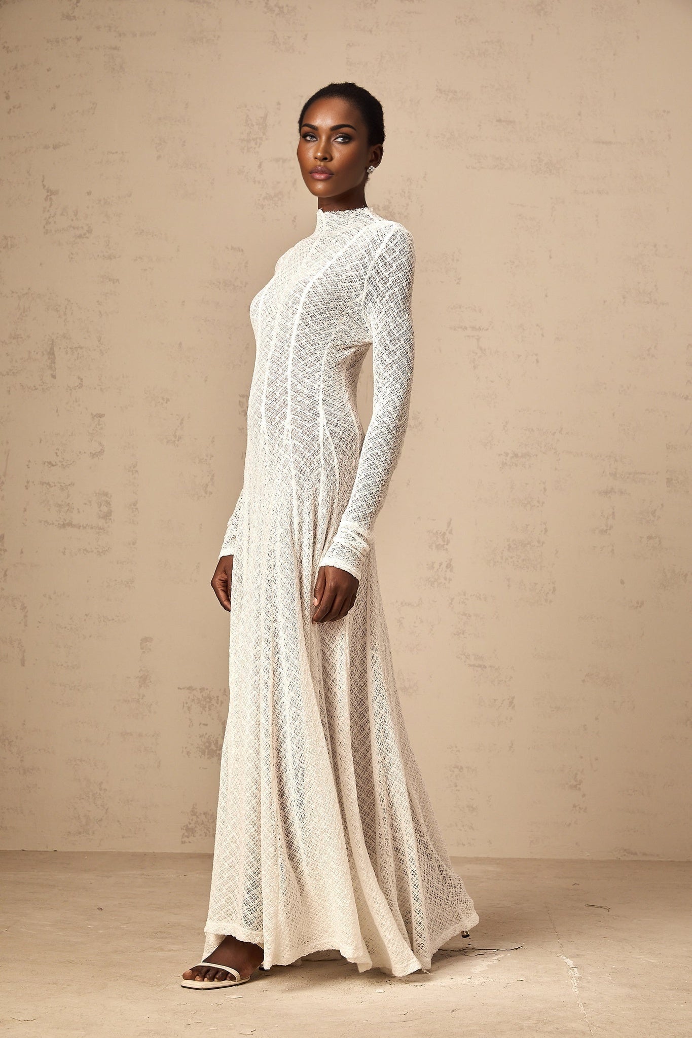 A long white lace maxi dress with a wavy pattern design worn by a model standing against a plain background