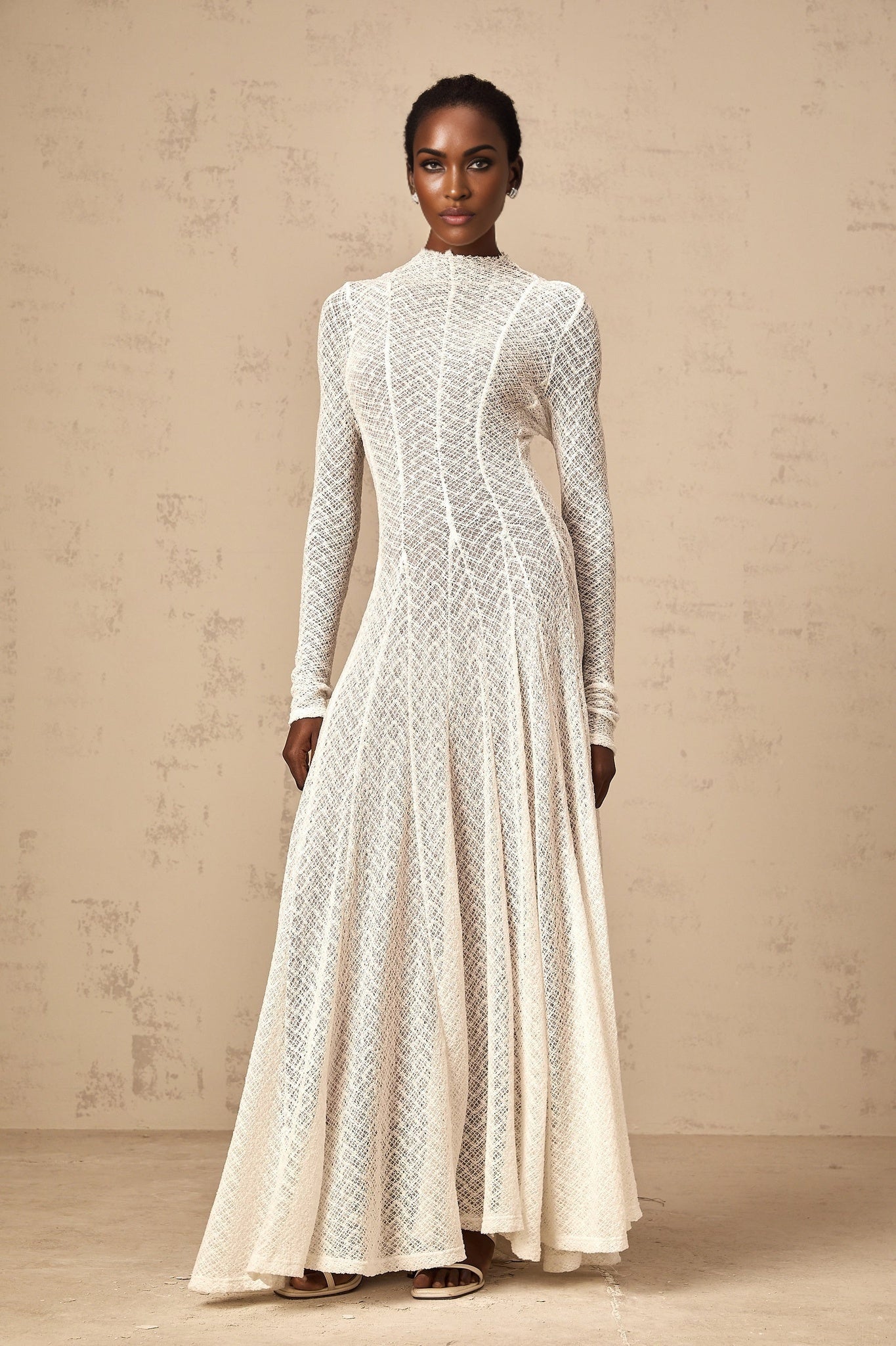 A long white lace maxi dress with a wavy pattern worn by a model standing against a plain background