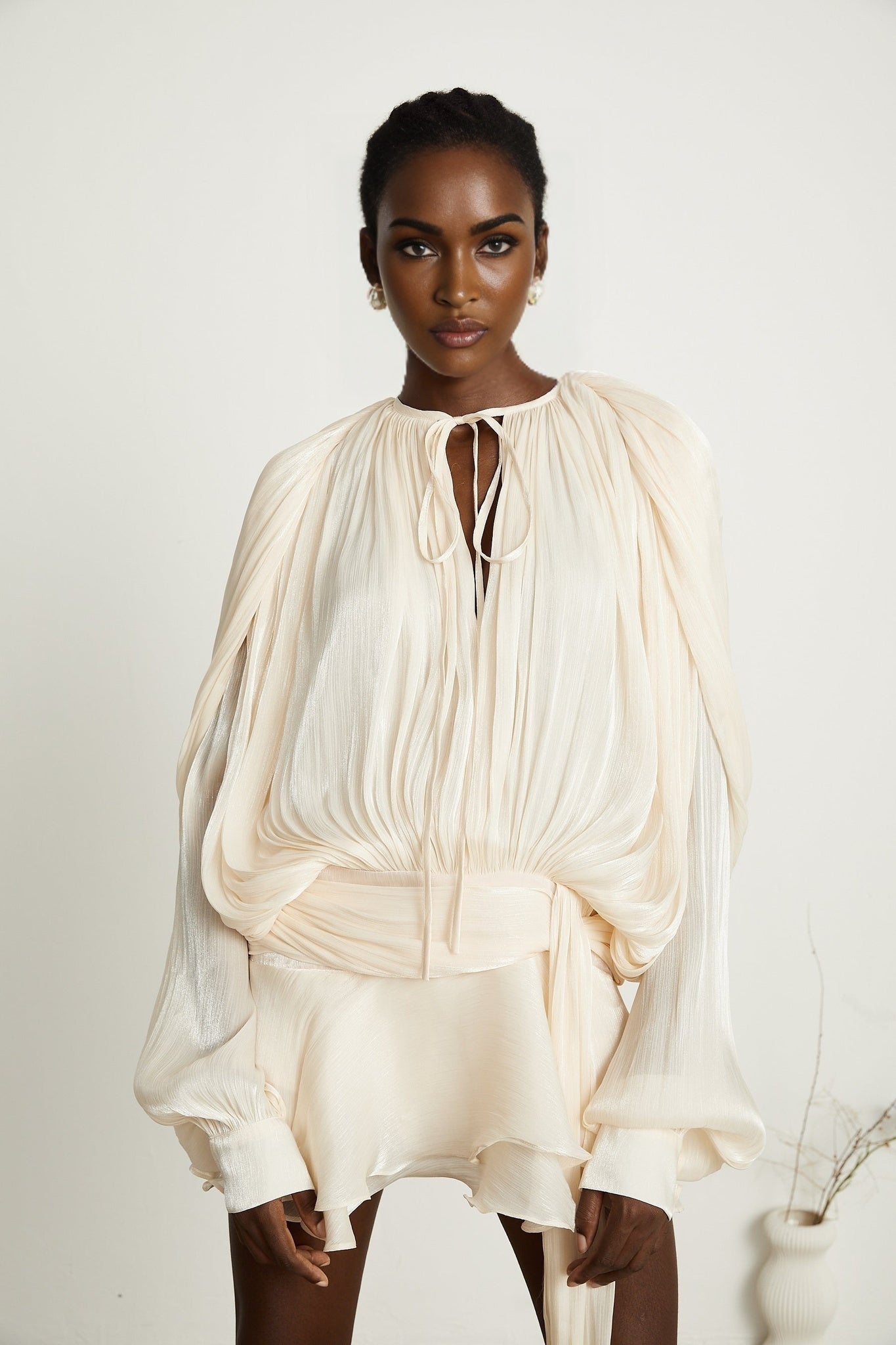 A neutralcolored mini dress with full pleats and ruffles featuring a short hemline and Eugenie style design