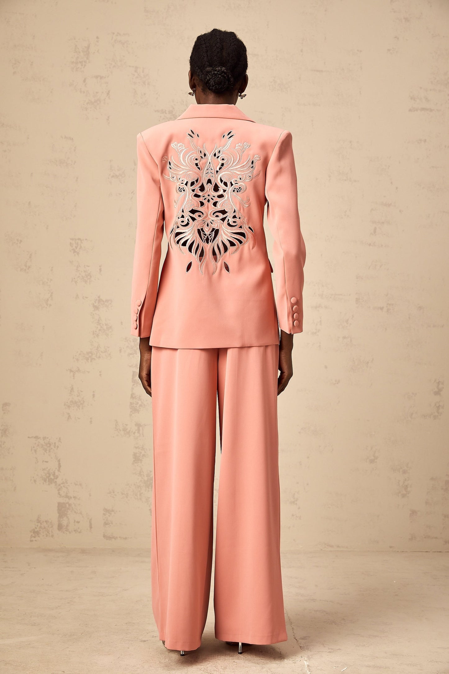 A pink blazer and trouser set with embroidery details and cutout designs The blazer features a tailored silhouette with decorative cutouts while the matching trousers have a sleek fit Both pieces are adorned with intricate pink embroidery creating an elegant and coordinated outfit