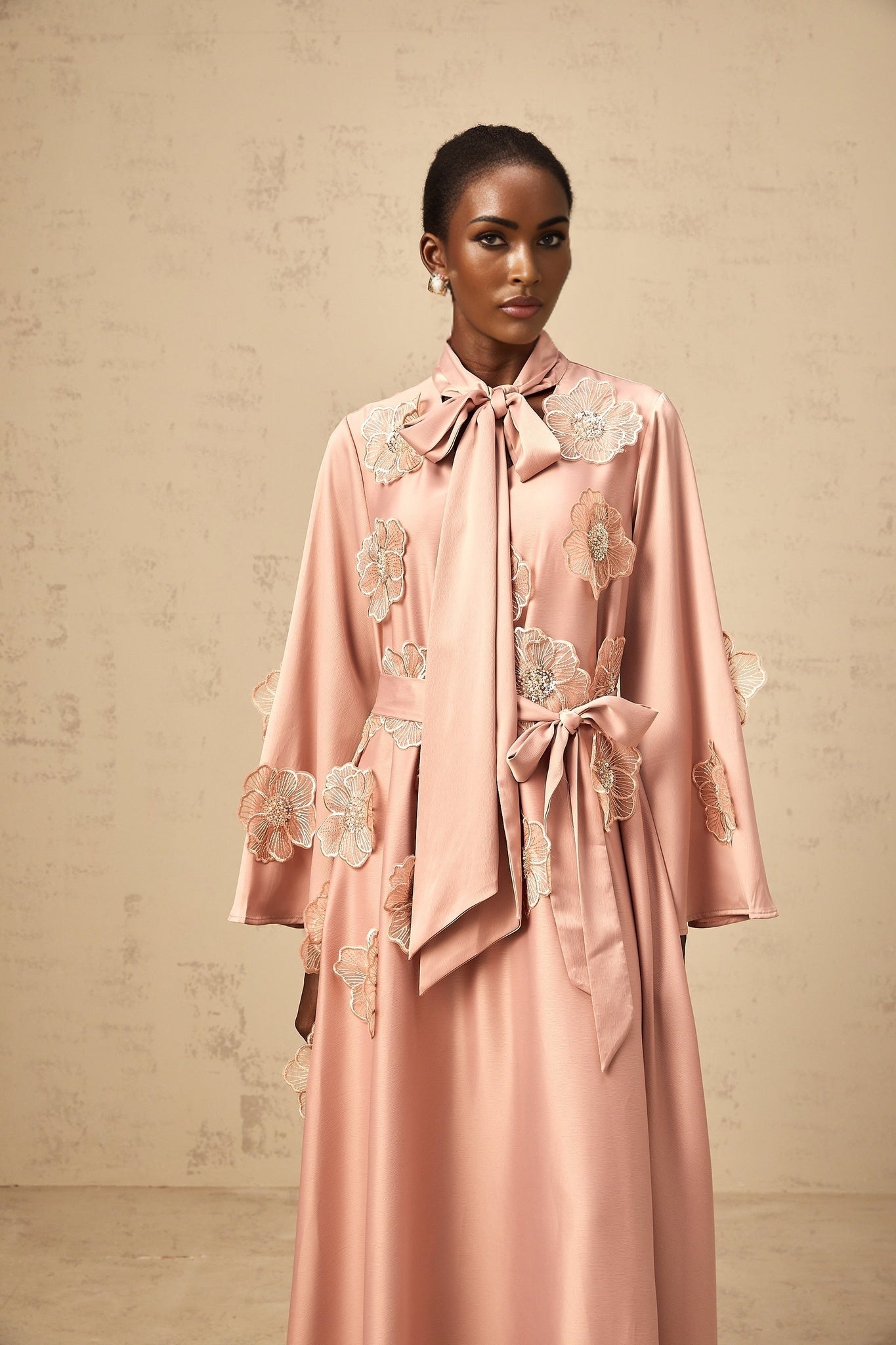 A pink flared midi dress with floral applique details and a tied waist design featuring a delicate Orane pattern
