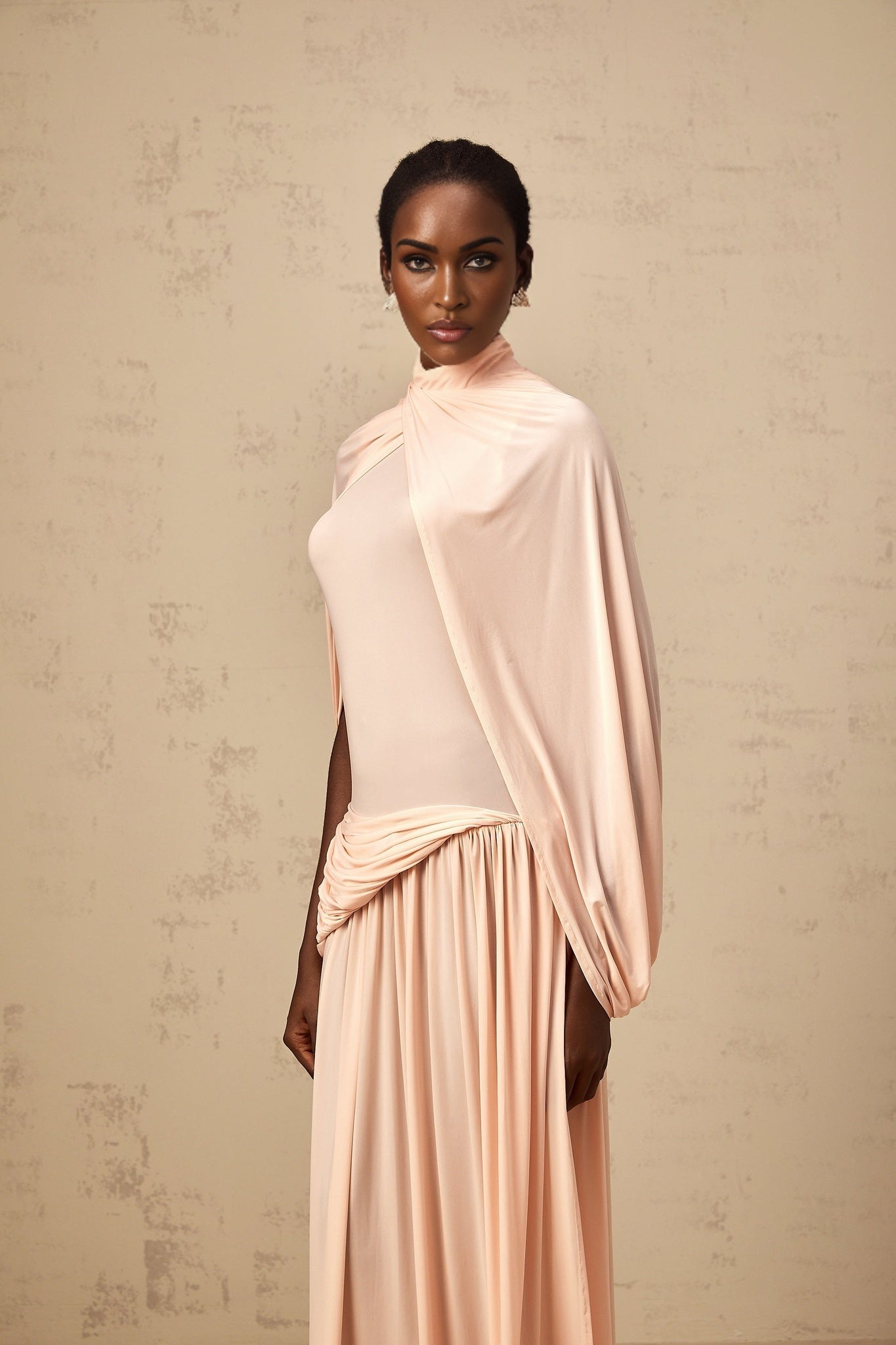 A pink maxi dress with a cape design and asymmetrical hemline worn by a model against a plain background