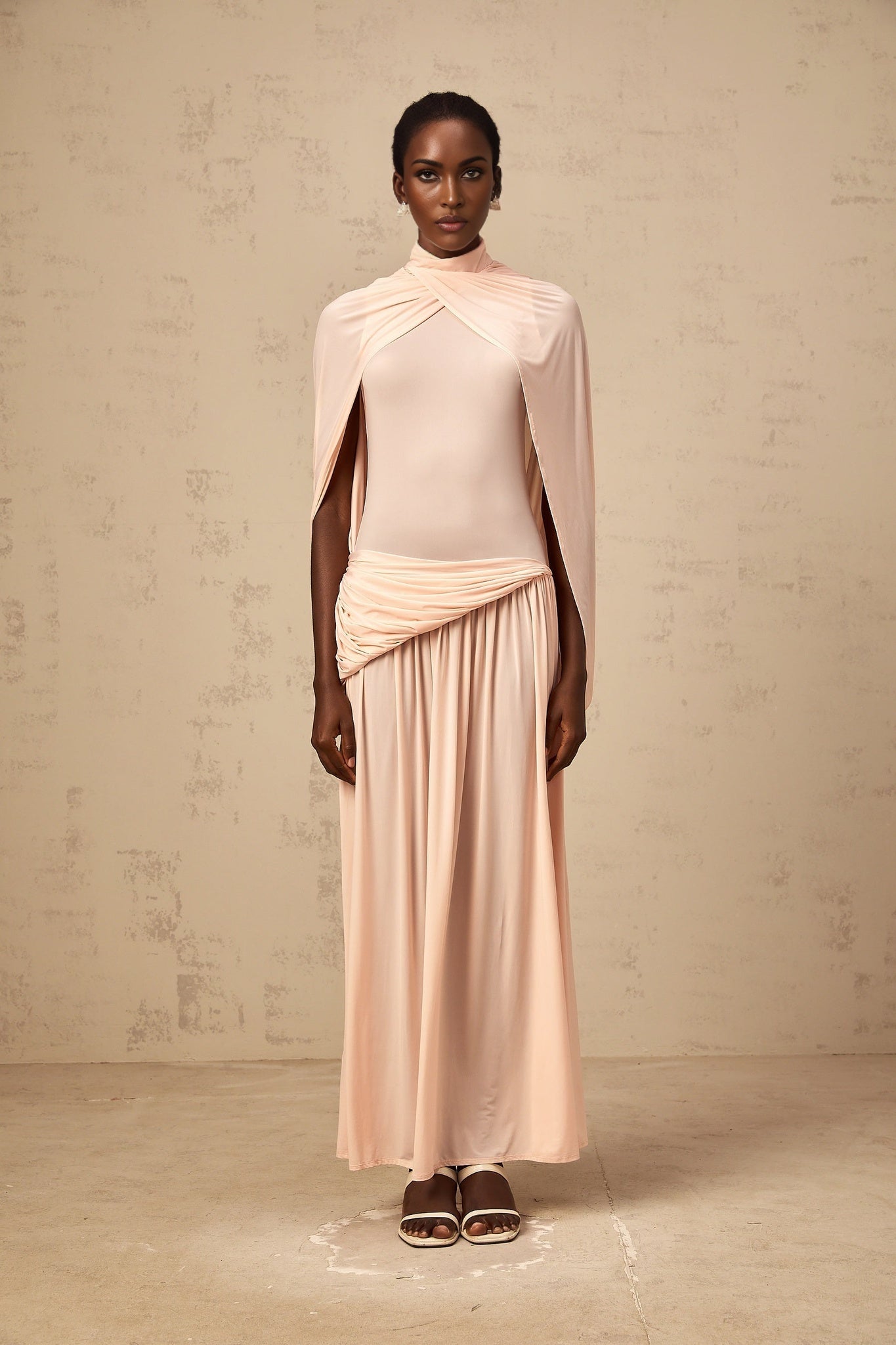 A pink maxi dress with a cape design and asymmetrical hemline worn by a model standing against a plain background