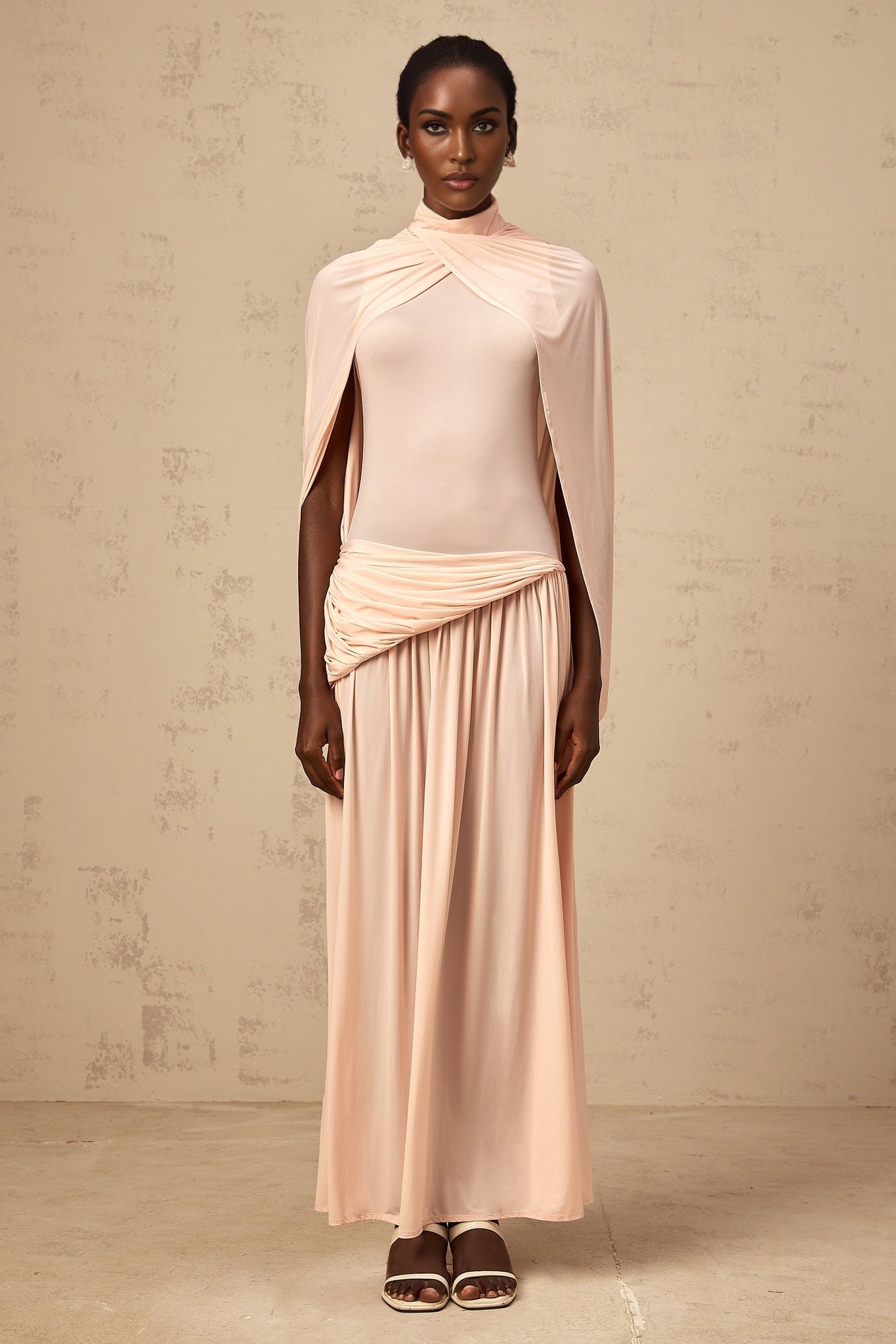 A pink maxi dress with an asymmetrical hemline and a capelike design over the shoulders featuring flowy fabric and a long silhouette