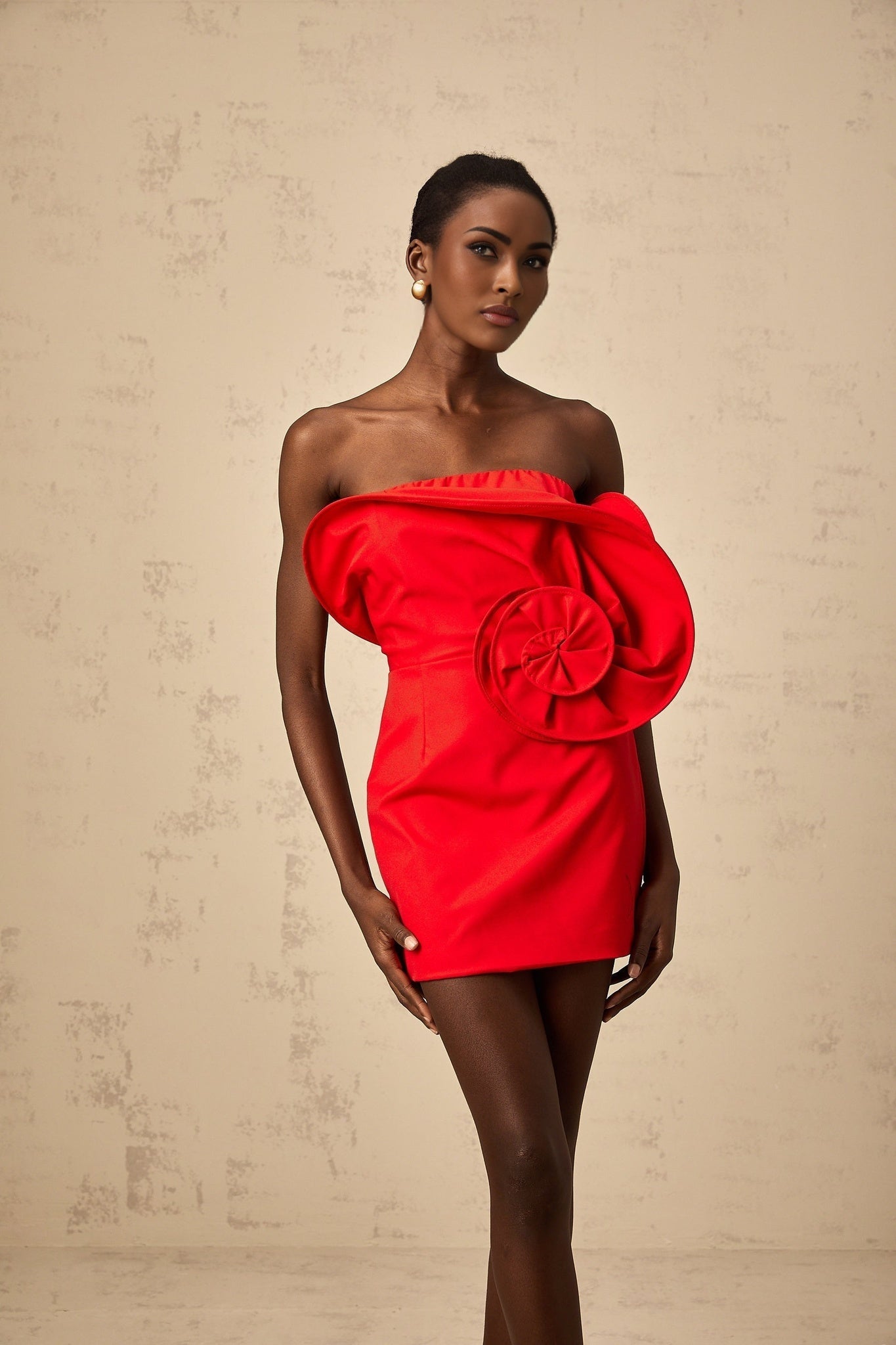 A red off shoulder mini dress with a sculptural rose detail on the bodice worn by a model against a plain background