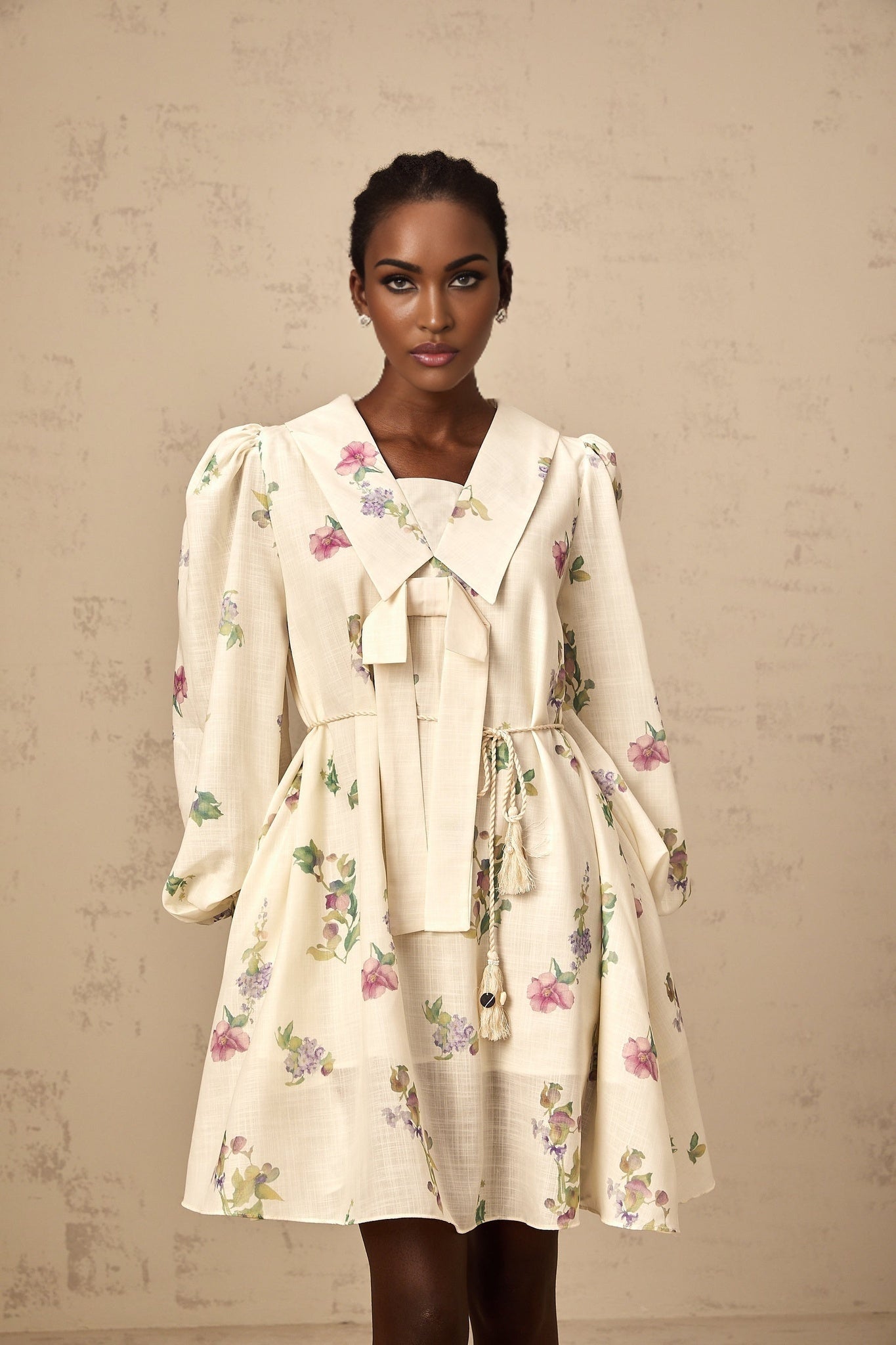 A short beige dress with a floral print pattern featuring a bow detail at the waist and a mini length skirt