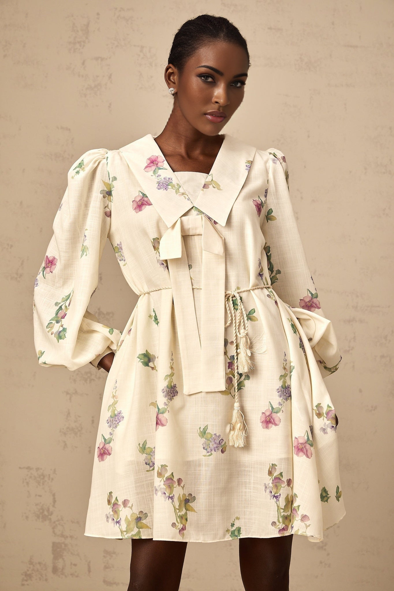 A short beige dress with a floral print pattern featuring a bow detail at the waist and a mini length skirt