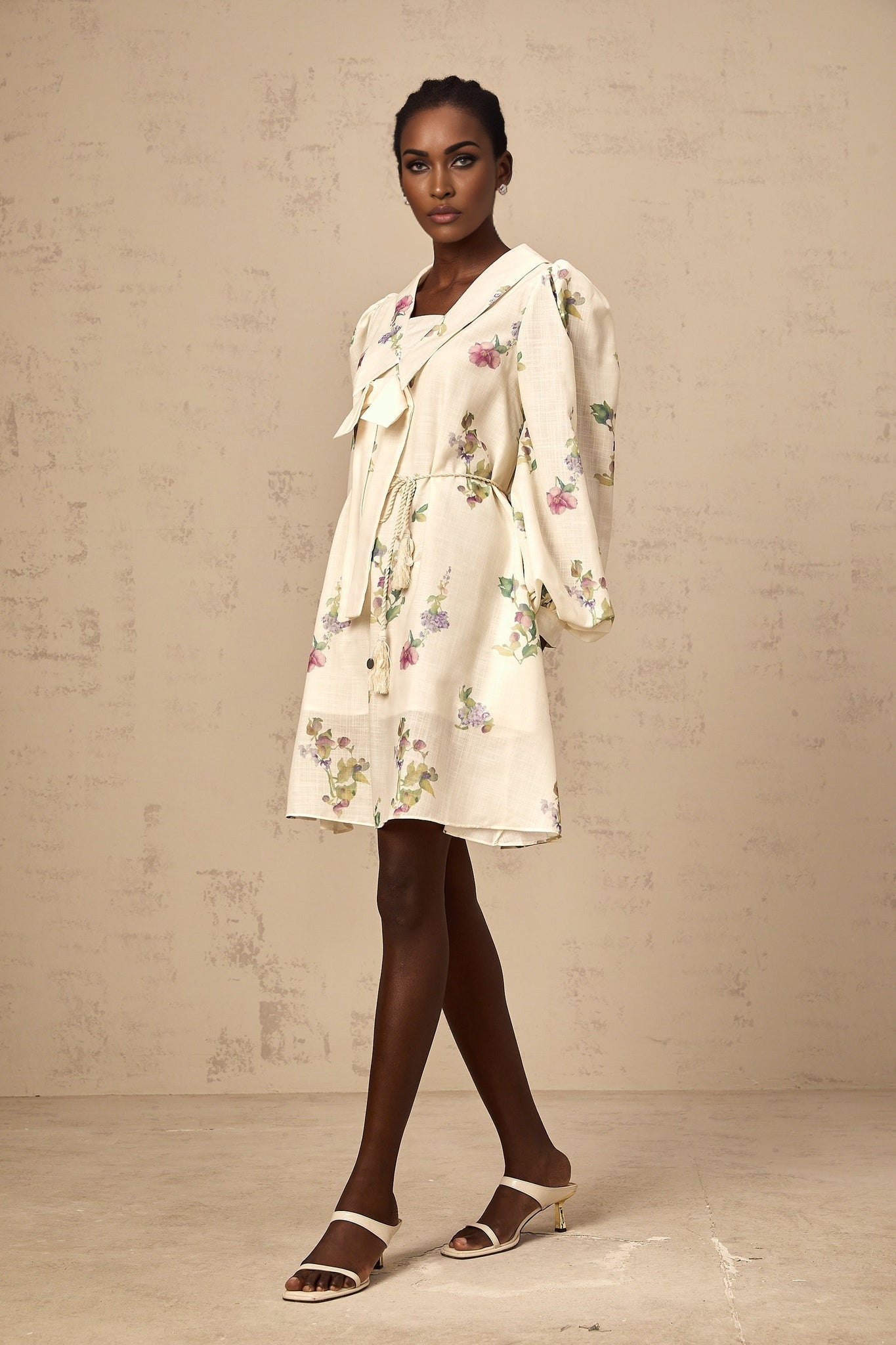 A short beige mini dress with a floral print pattern featuring a bow detail at the waist and a flowy skirt