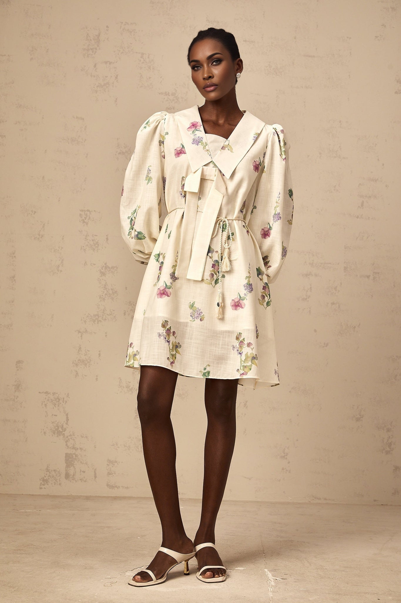 A short beige mini dress with a floral print pattern featuring a large bow tied at the front neckline and short sleeves
