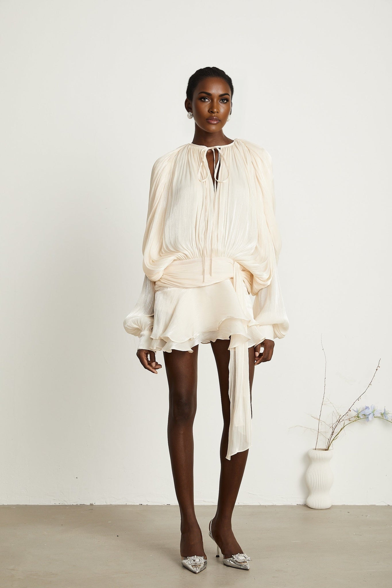 A short neutralcolored mini dress with full pleats and ruffles featuring a design inspired by Eugenie style