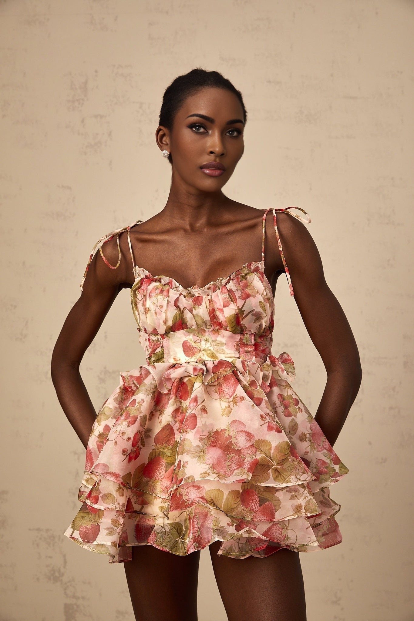 A short pink dress with a strawberry print pattern featuring a tiered skirt and likely short sleeves or straps styled in a feminine and playful design