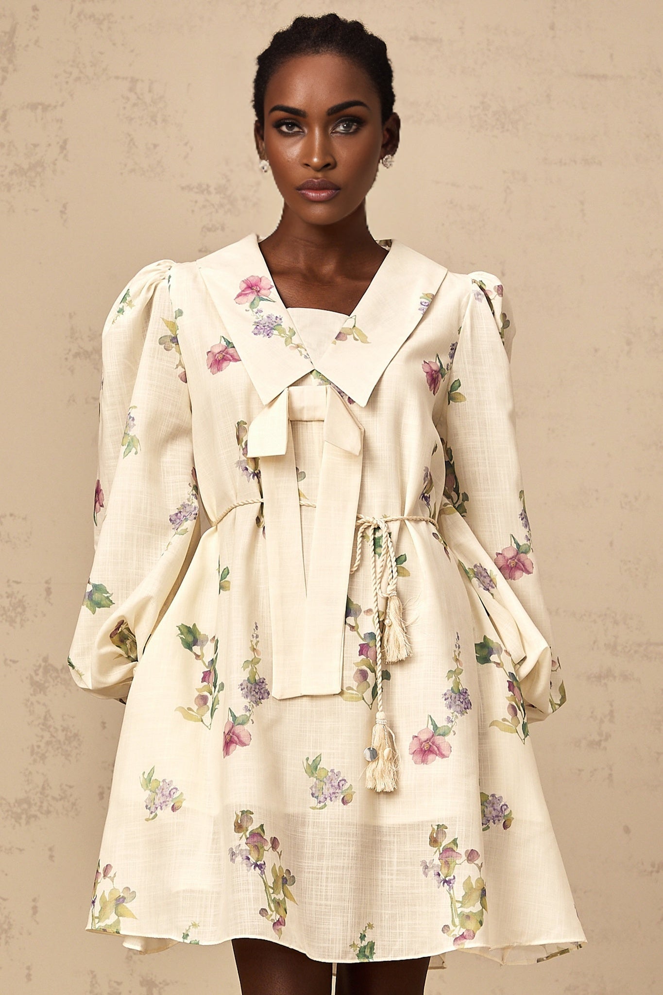 A short sleeveless mini dress in neutral tones featuring a floral print pattern and a decorative bow detail at the waist