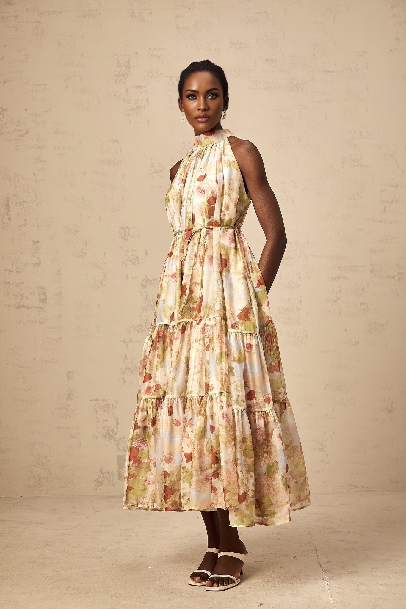 A sleeveless midi dress with a floral print in neutral colors worn by a model standing in a relaxed pose