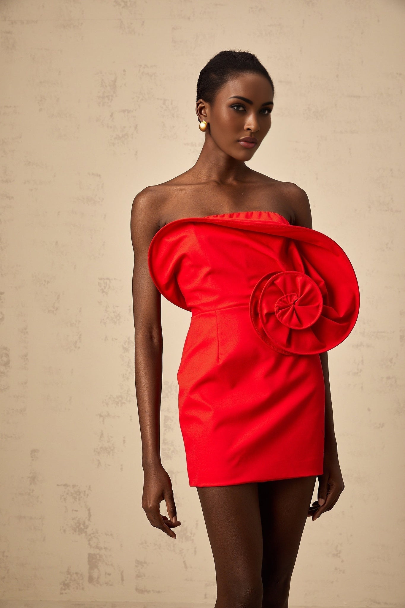 A vibrant red mini dress with an offshoulder neckline featuring a large sculptural rose detail on the bodice creating a dramatic and romantic silhouette