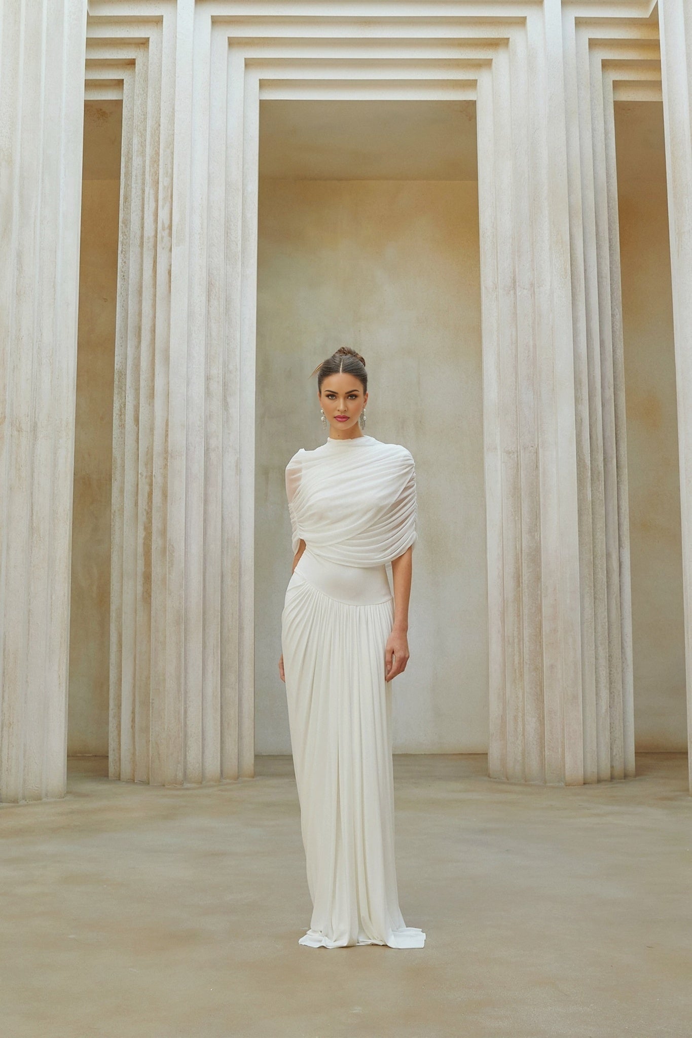 A white asymmetrical maxi dress with pleated details worn by a model against a plain background