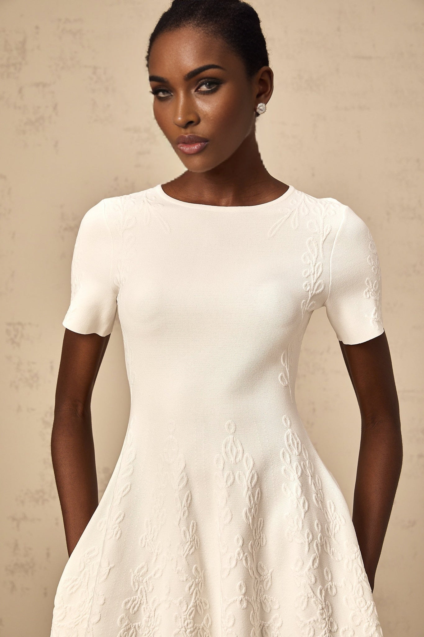 A white knitted mini dress with cut out details and jacquard pattern worn by a model standing against a plain background