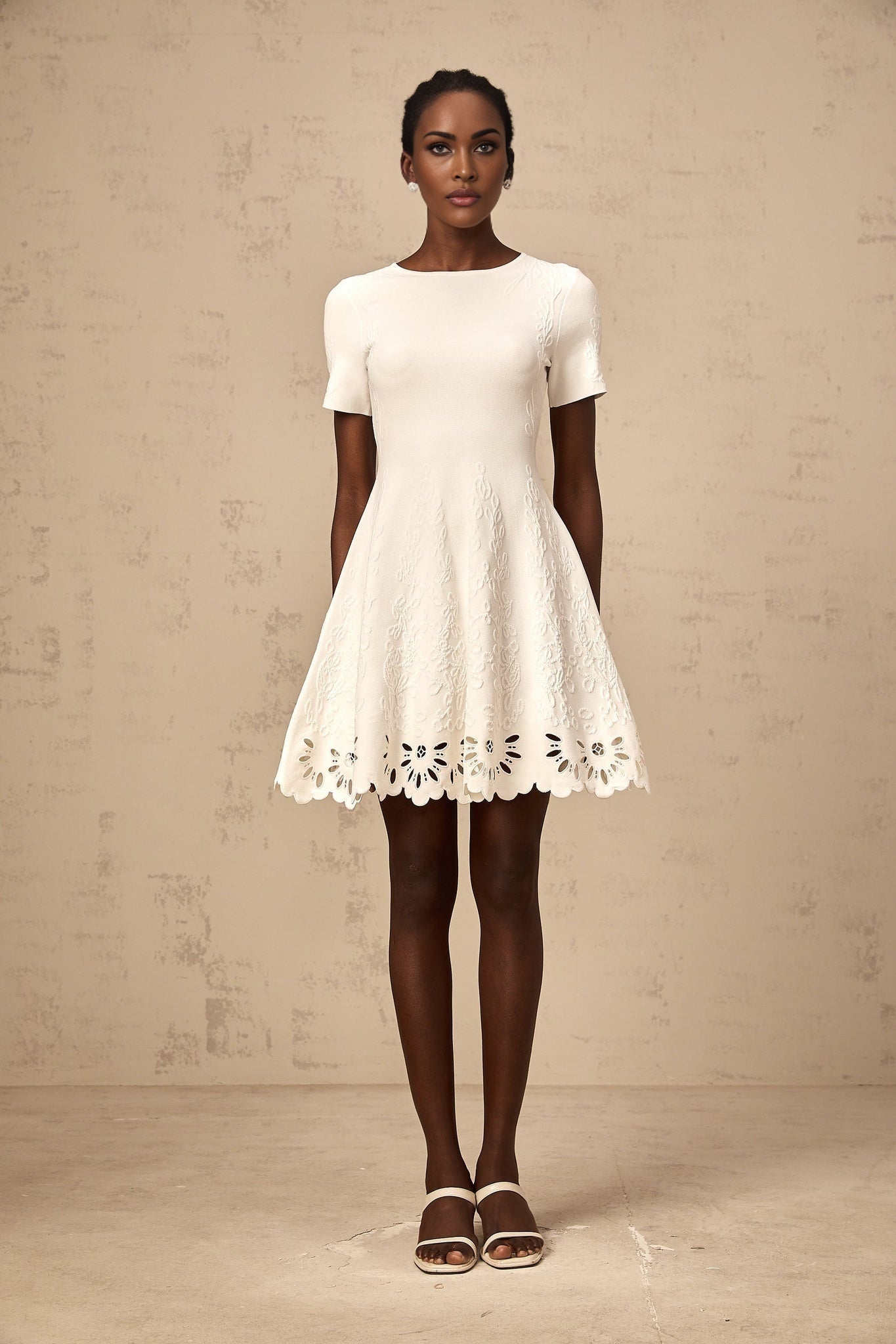 A white knitted mini dress with cutout jacquard pattern worn by a model standing against a plain background