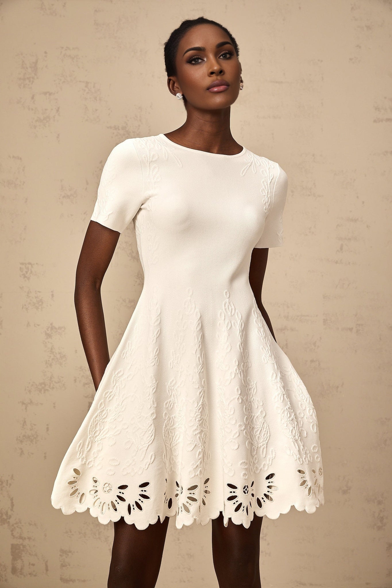 A white knitted mini dress with jacquard pattern and cutout details modeled by a woman named Emilie