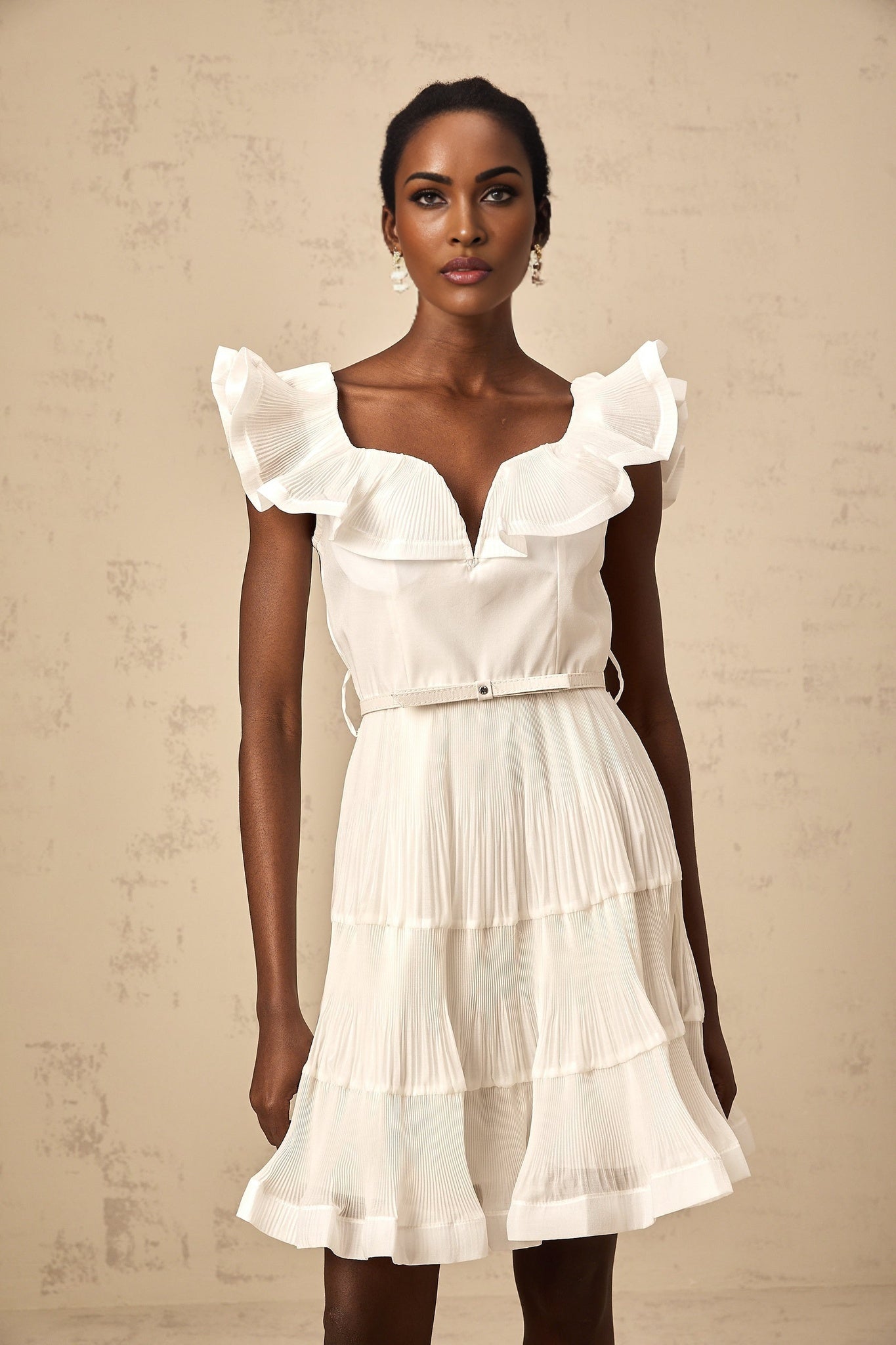 A white mini dress with a frilled neckline and pleated skirt worn by a model against a plain background