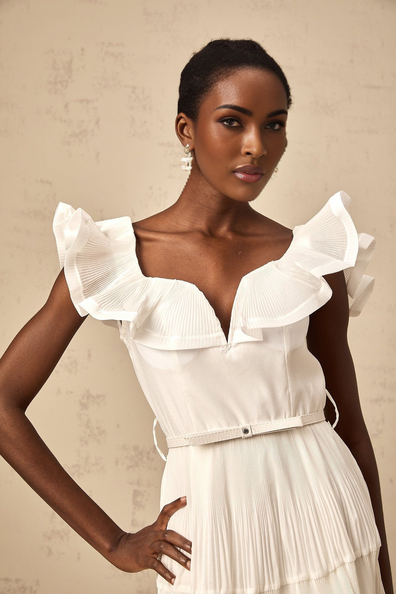 A white mini dress with a frilled neckline and pleated skirt worn by a model standing against a plain background